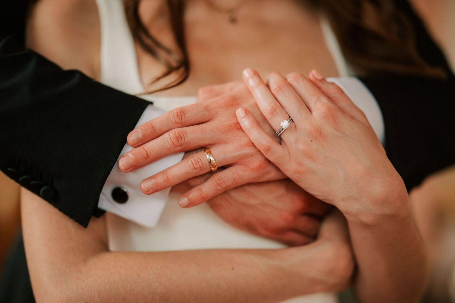 Close-up of couple's hands showing wedding and engagement rings, bride in white dress, groom in black suit.