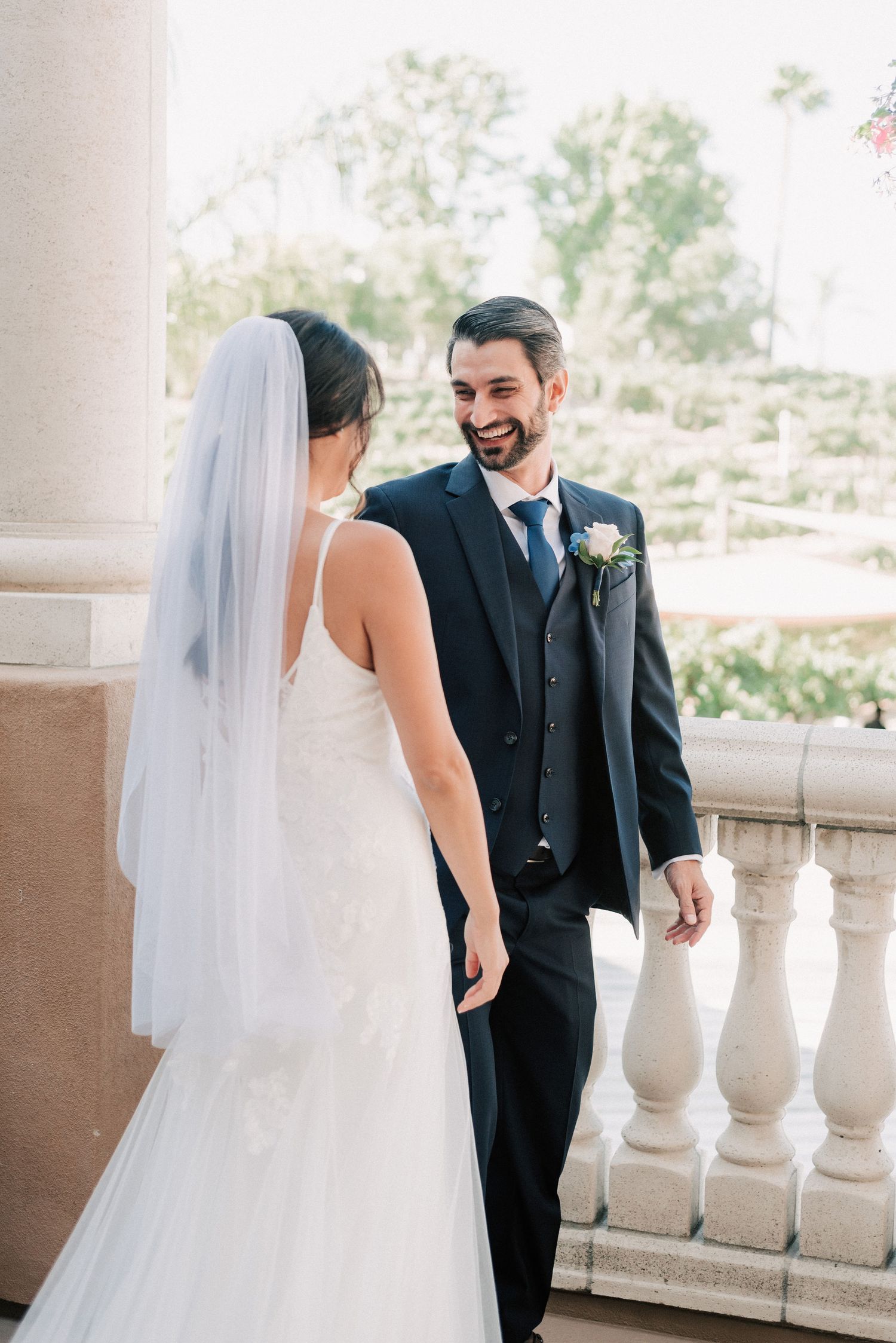 Bride in white gown and veil shares a joyful first look moment with groom in navy suit on elegant balcony.