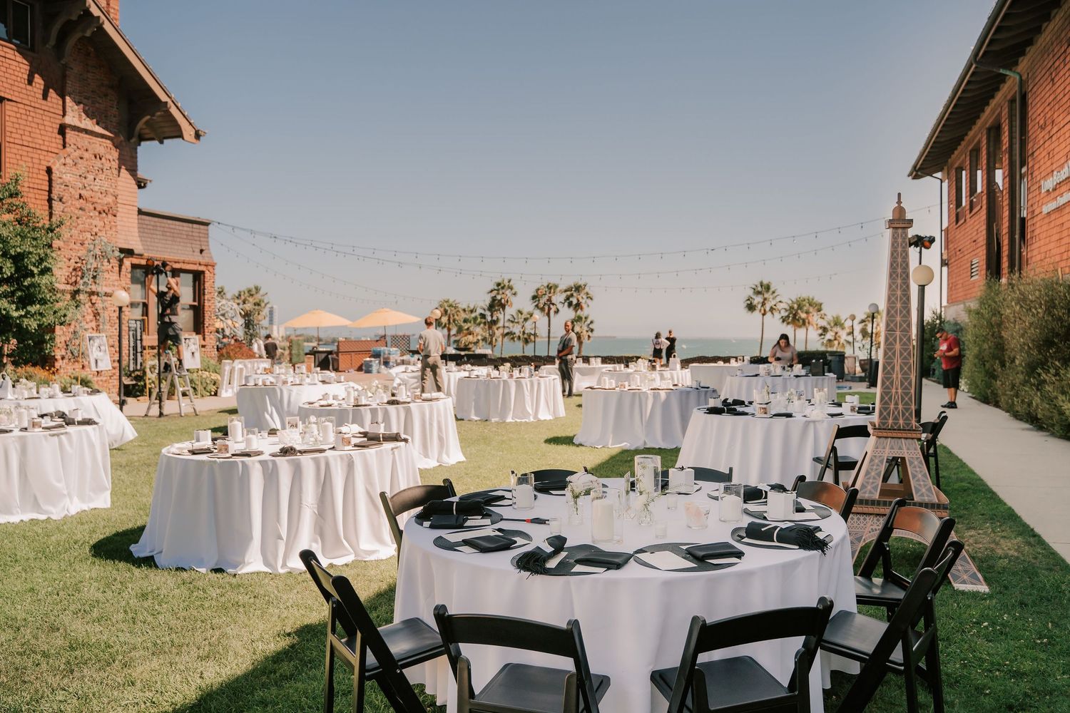 Outdoor wedding reception on a sunny lawn with round tables set with white linens, black chairs, palm trees, and a brick building nearby.