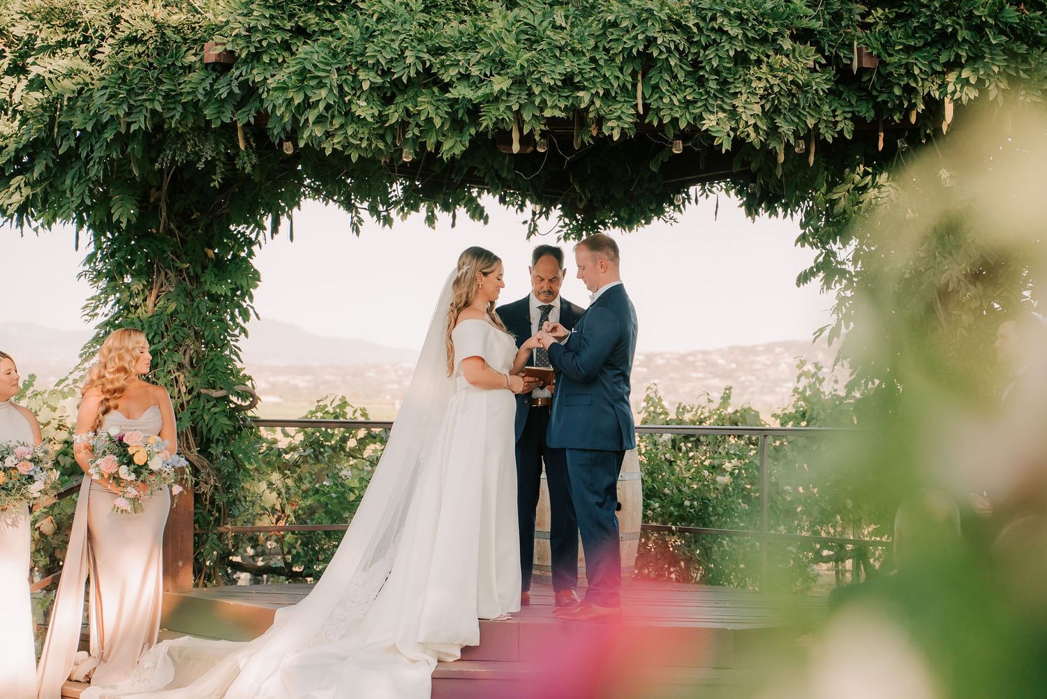 Bride and groom exchange vows under lush green arbor at outdoor wedding ceremony with scenic mountain views.