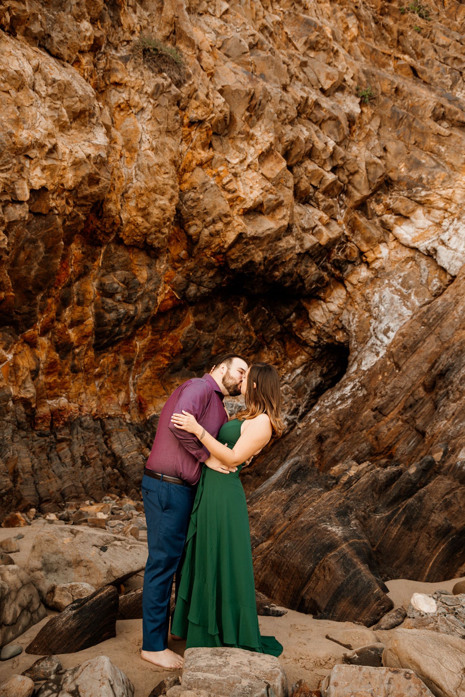 Couple embracing romantically in front of dramatic rocky cliffs, woman in green gown and man in burgundy shirt.