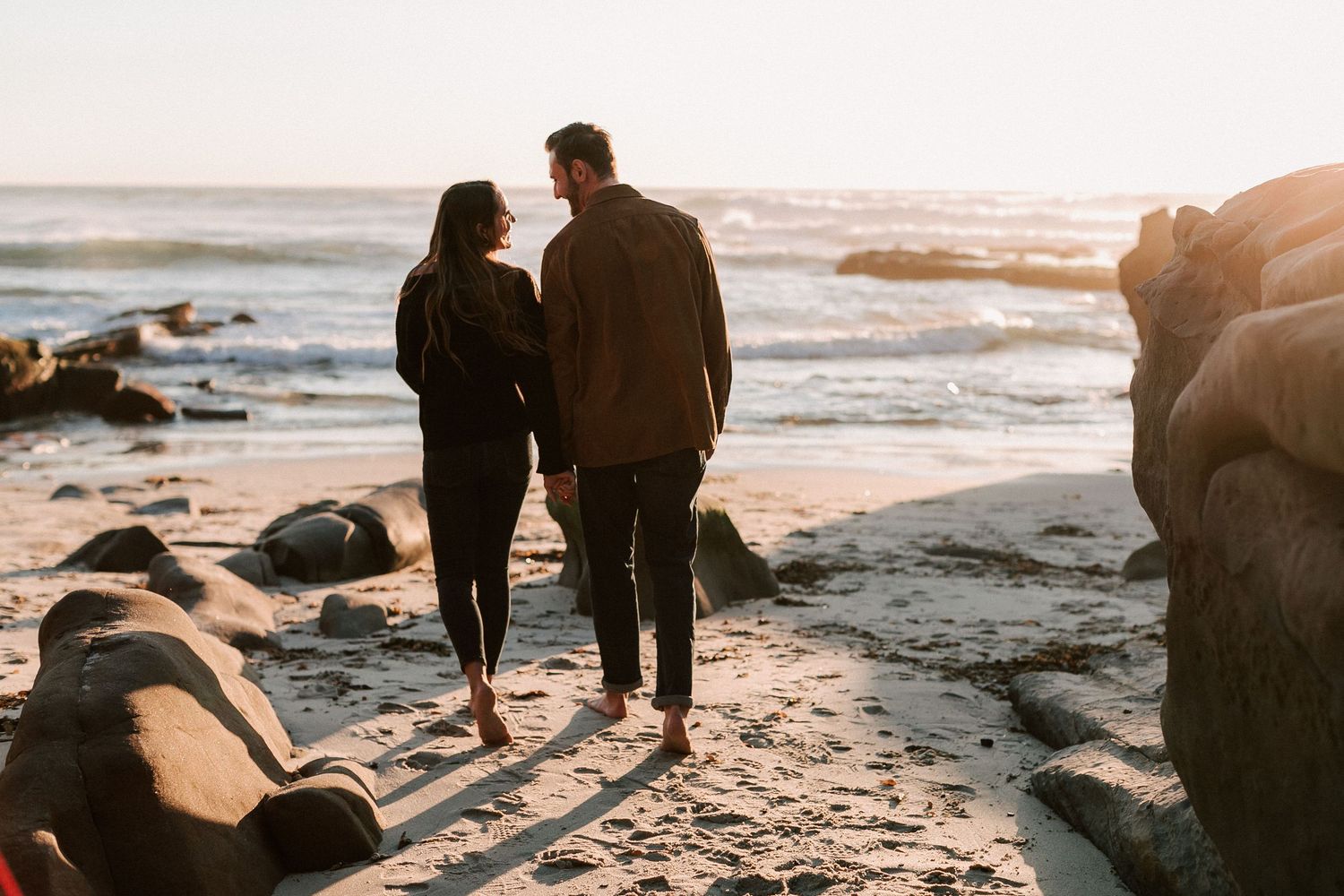 Couple walking arm in arm along a sandy beach at sunset with rocky outcroppings nearby.