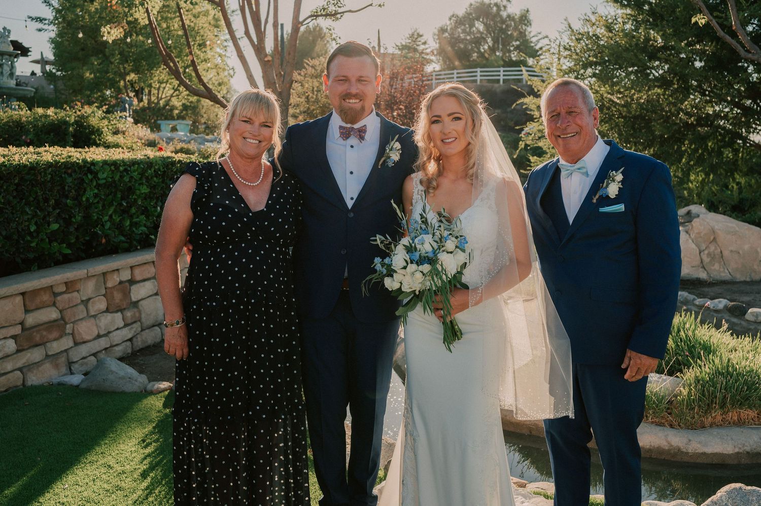 Wedding party of four posing outdoors, bride in white gown with bouquet, groom and guests in formal attire.