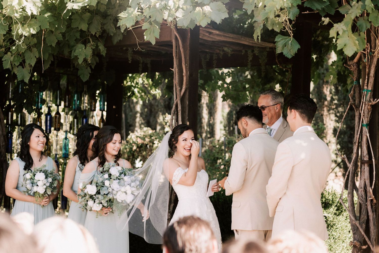 Bride smiling at groom during outdoor wedding ceremony under lush green vine arbor with bridesmaids in gray dresses.