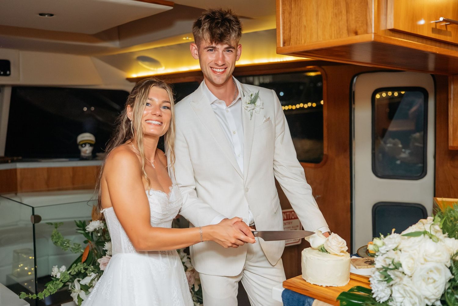 Smiling bride and groom cut white wedding cake together aboard a yacht at their reception.