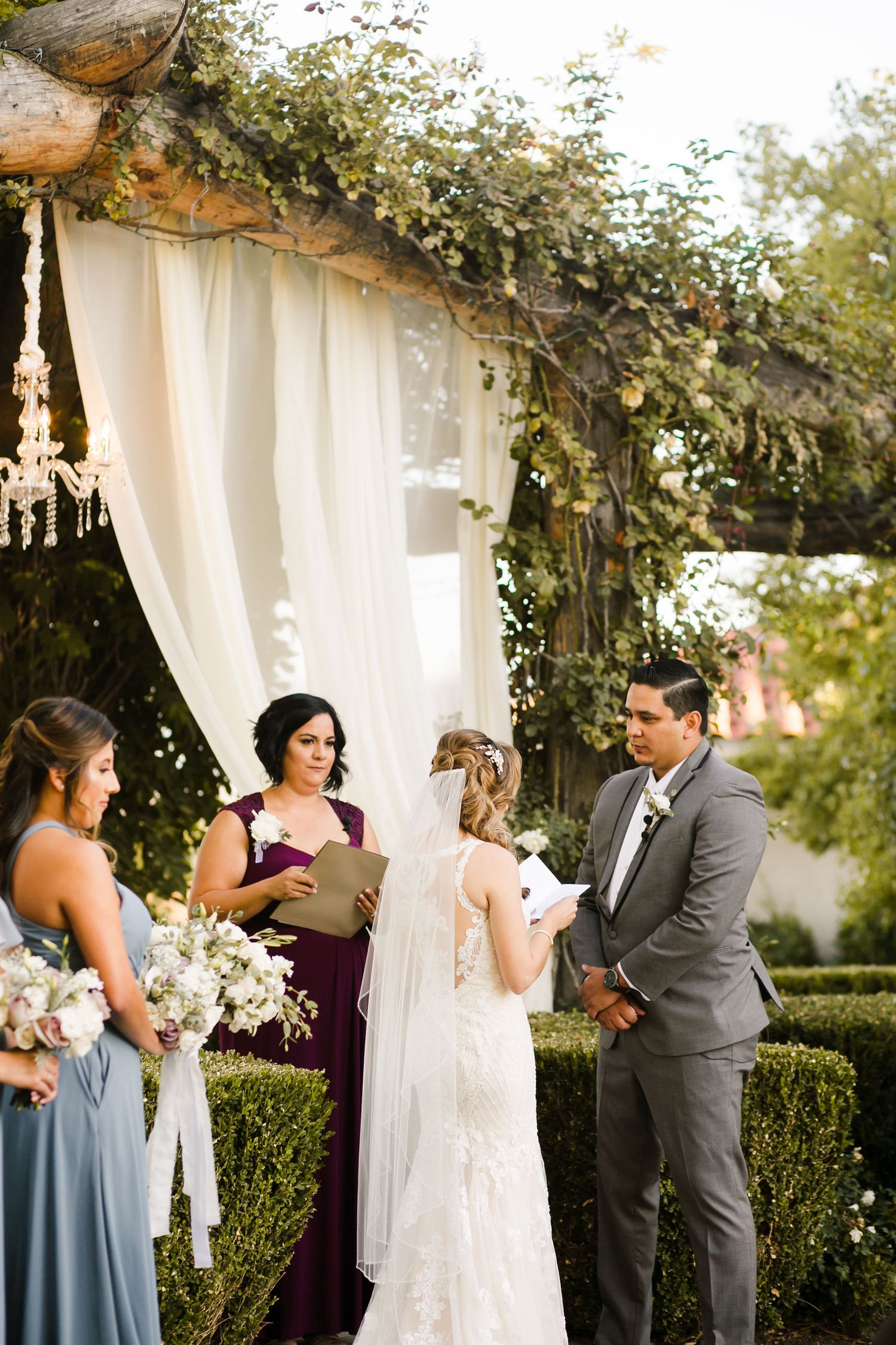 Outdoor wedding ceremony with bride and groom exchanging vows under a rustic floral arch with white draping and chandeliers.