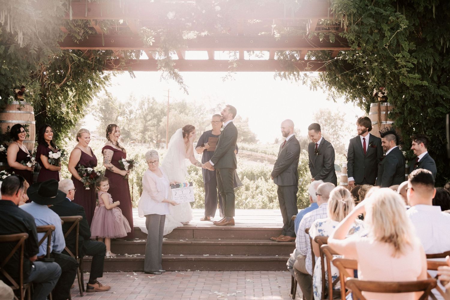 Outdoor wedding ceremony under a vine-covered pergola with bridesmaids in burgundy dresses and guests seated on wooden chairs.