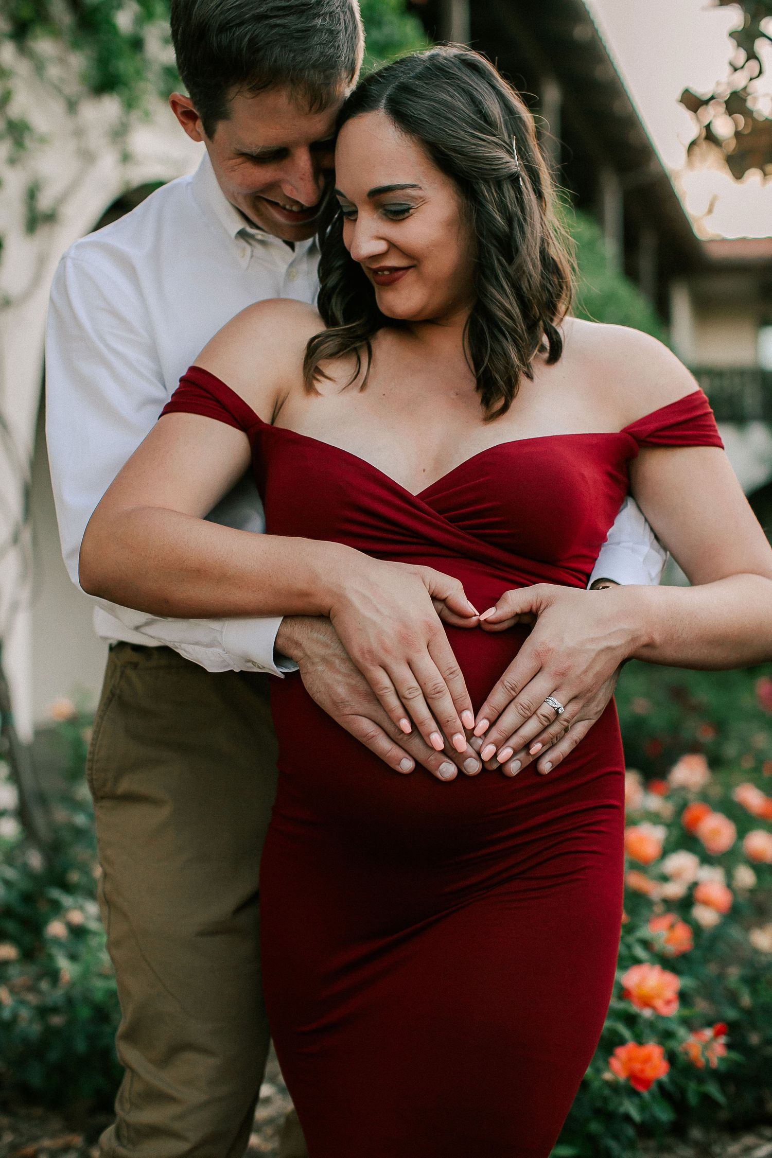 Couple forming heart shape on pregnant belly in red maternity dress among garden flowers.