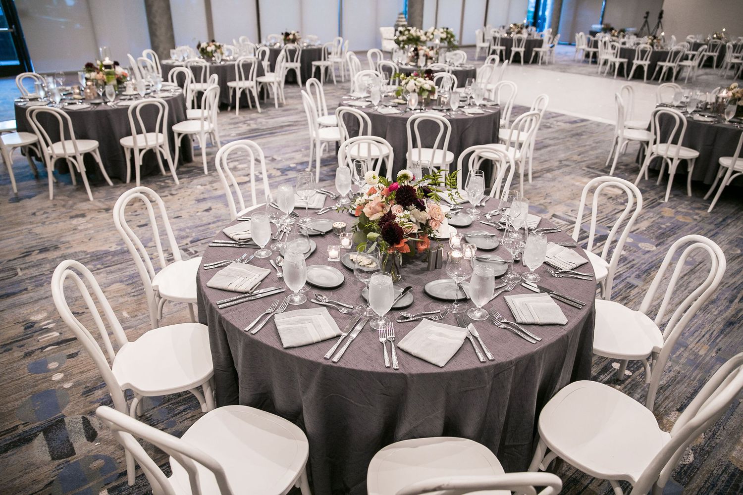 Elegant wedding reception hall with round tables, gray linens, white bentwood chairs, and floral centerpieces in a modern ballroom.