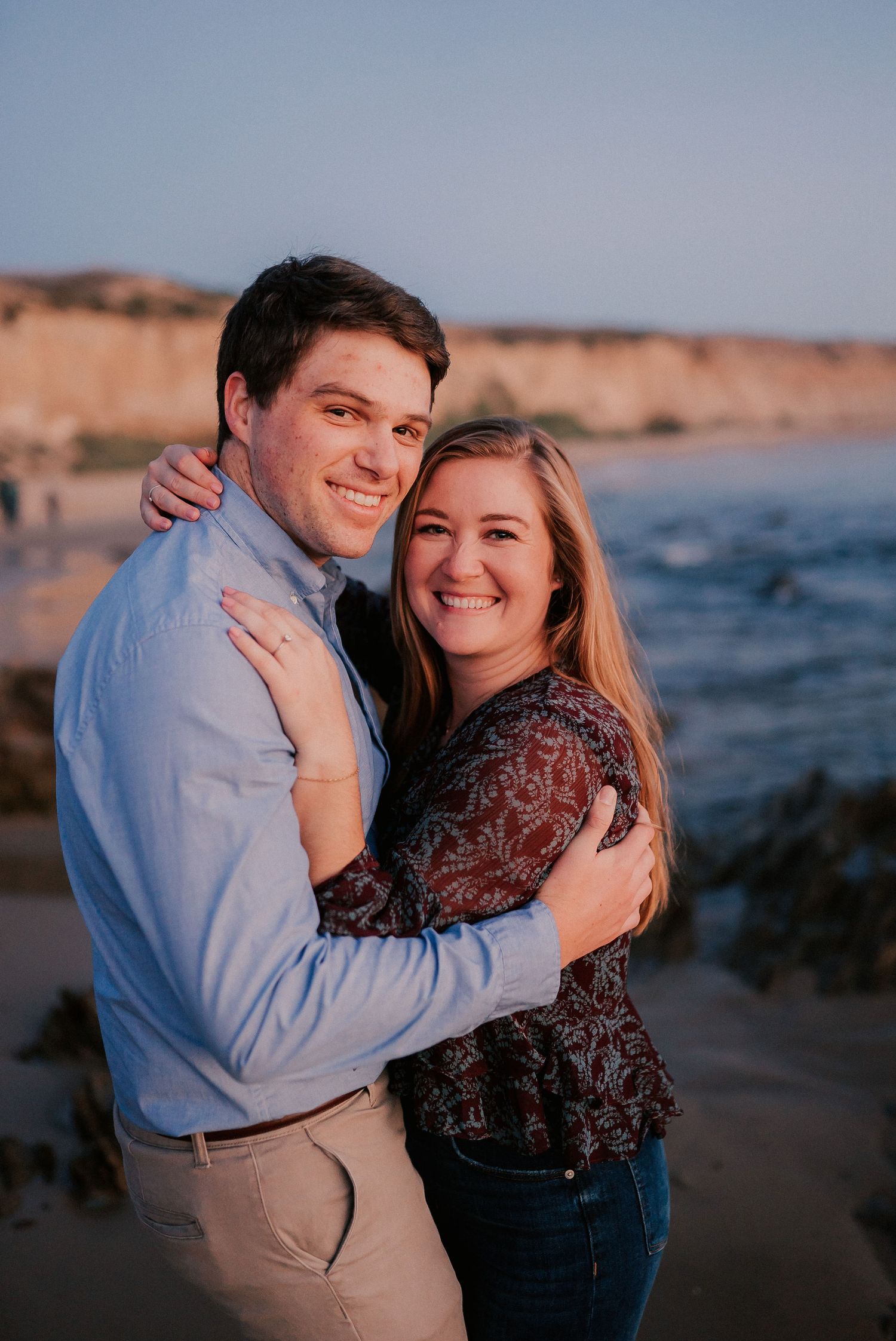 Smiling couple embracing on sunny beach with golden cliffs and ocean in background.