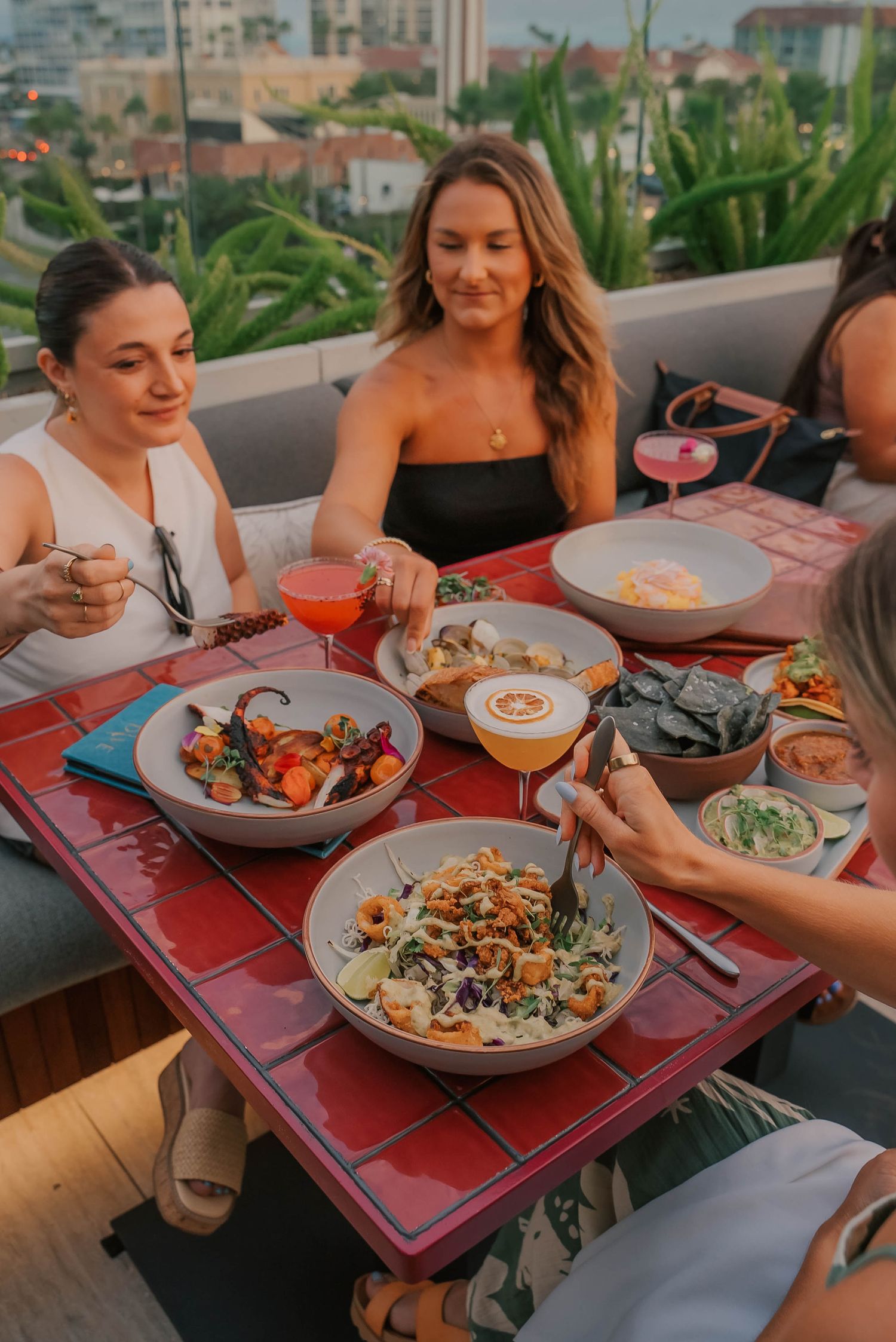 Women dining outdoors on rooftop with colorful dishes, cocktails, and city views at sunset.
