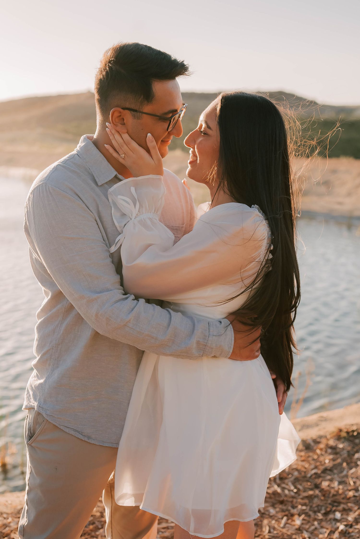 Couple sharing a romantic moment by a sunlit lake, woman in white dress and man in gray shirt embracing outdoors.