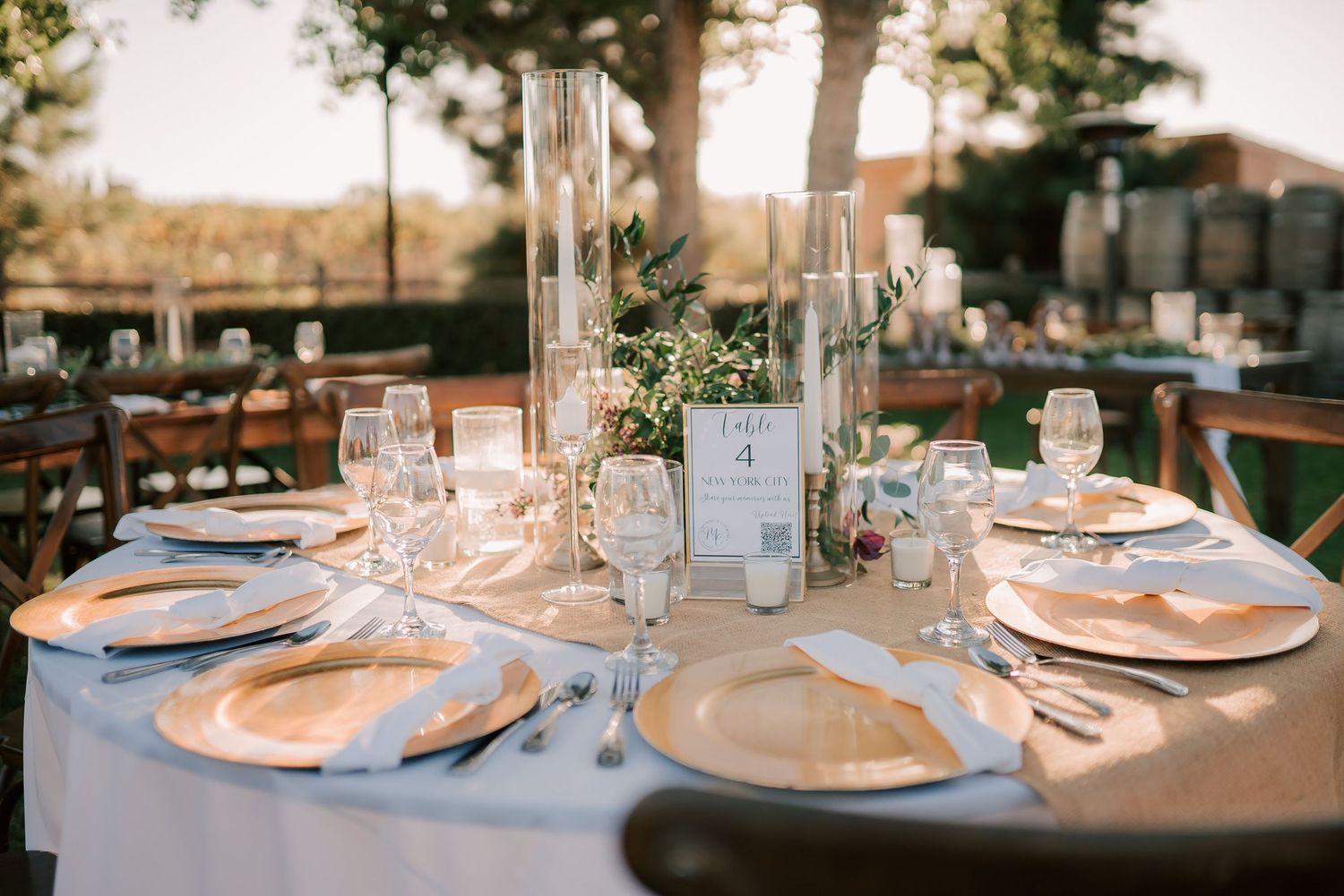 Elegant outdoor wedding reception table set with gold chargers, crystal glasses, and floral centerpieces on a light blue tablecloth.