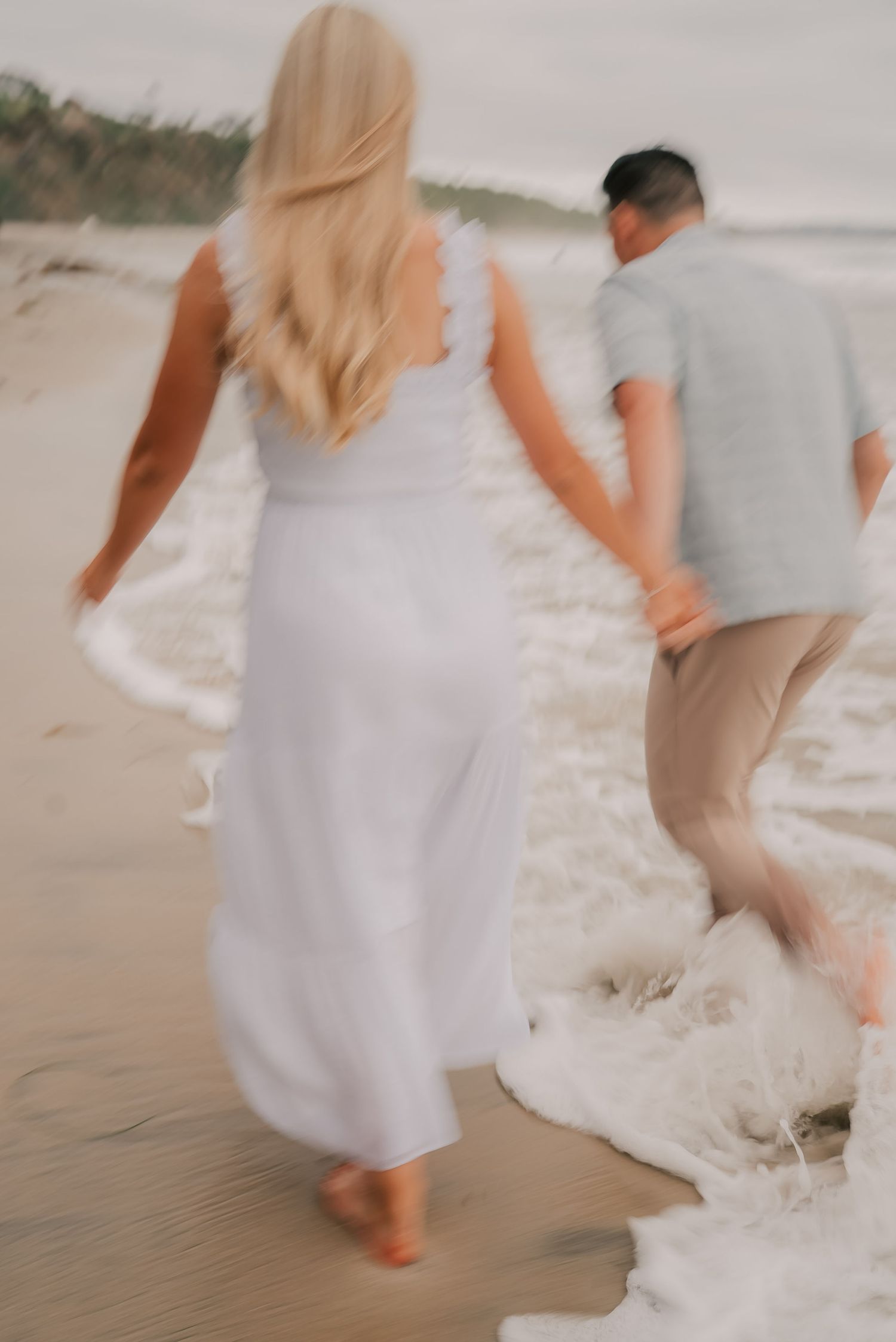 Blurry romantic photo of a couple holding hands walking along a sandy beach shore in soft pastel outfits.