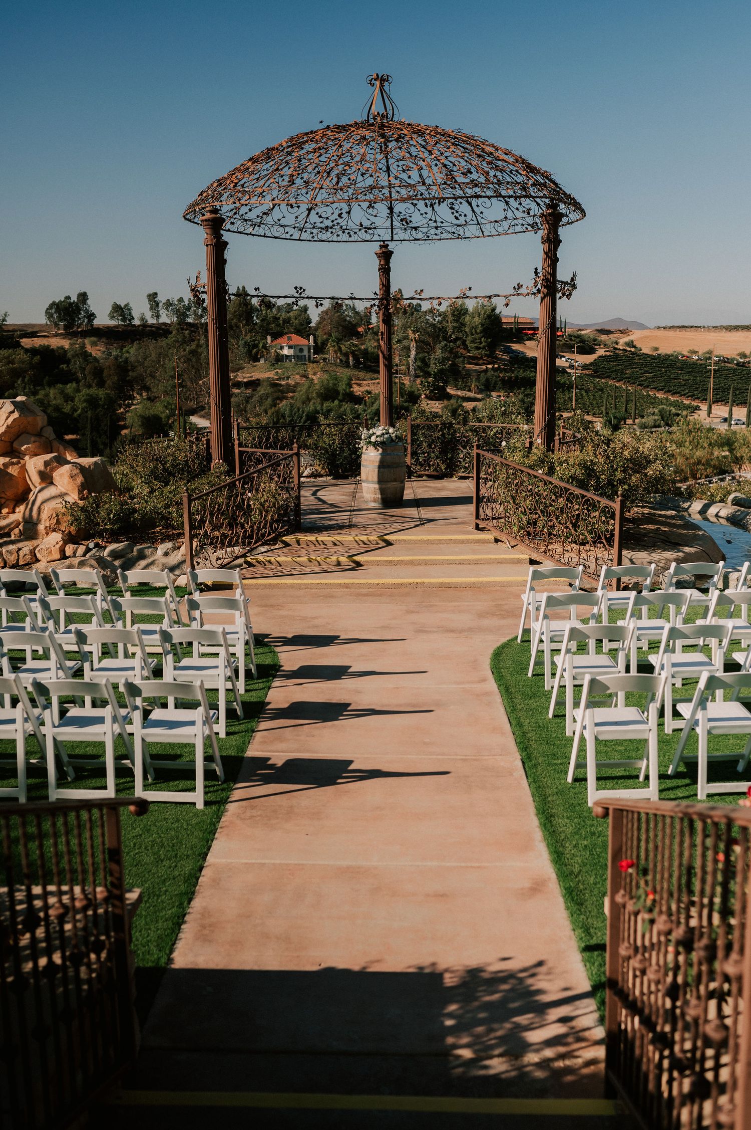 Elegant outdoor wedding ceremony setup with white chairs, a rustic iron gazebo altar, and scenic vineyard views.