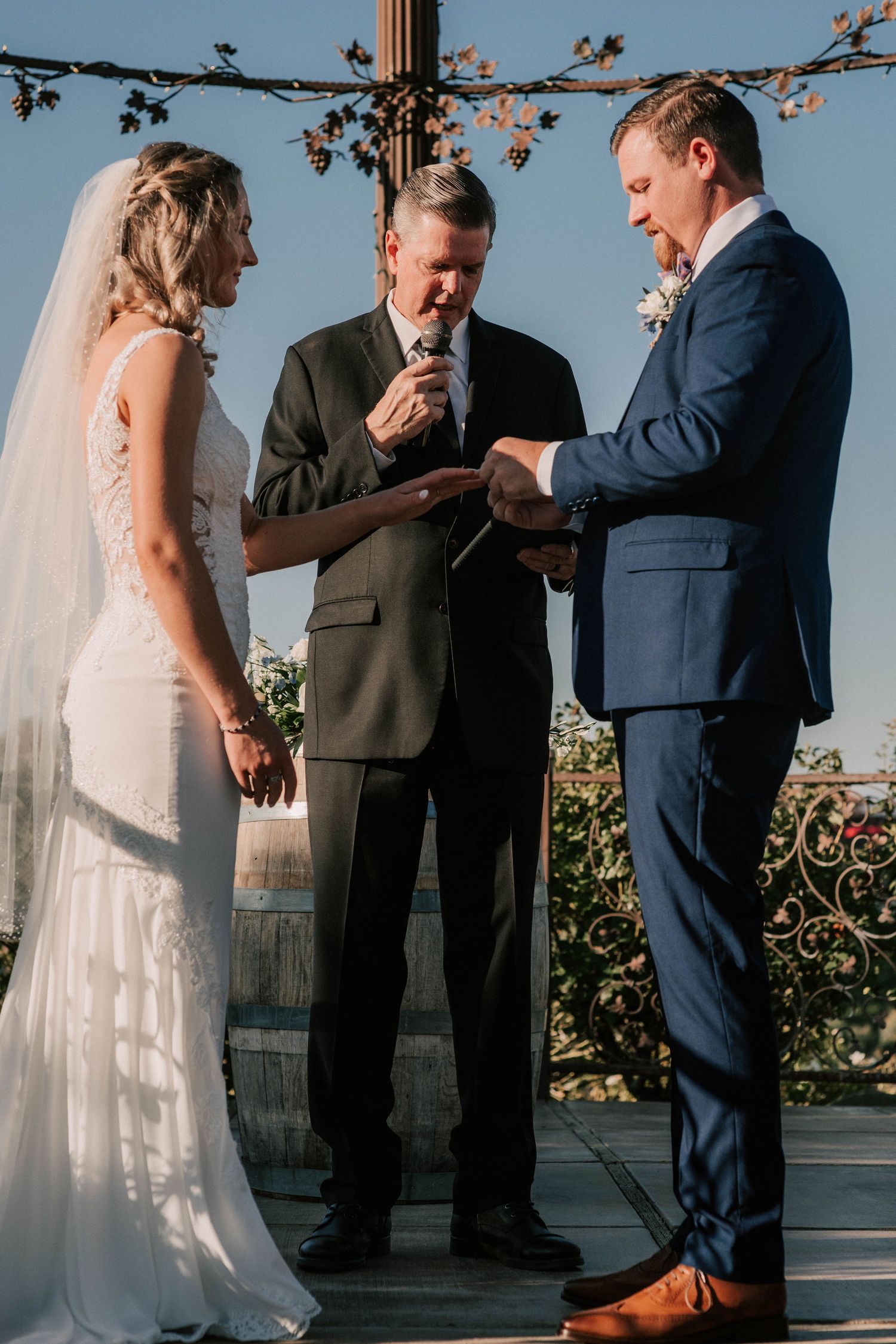 Bride and groom exchange rings during outdoor wedding ceremony with officiant at a vineyard venue.