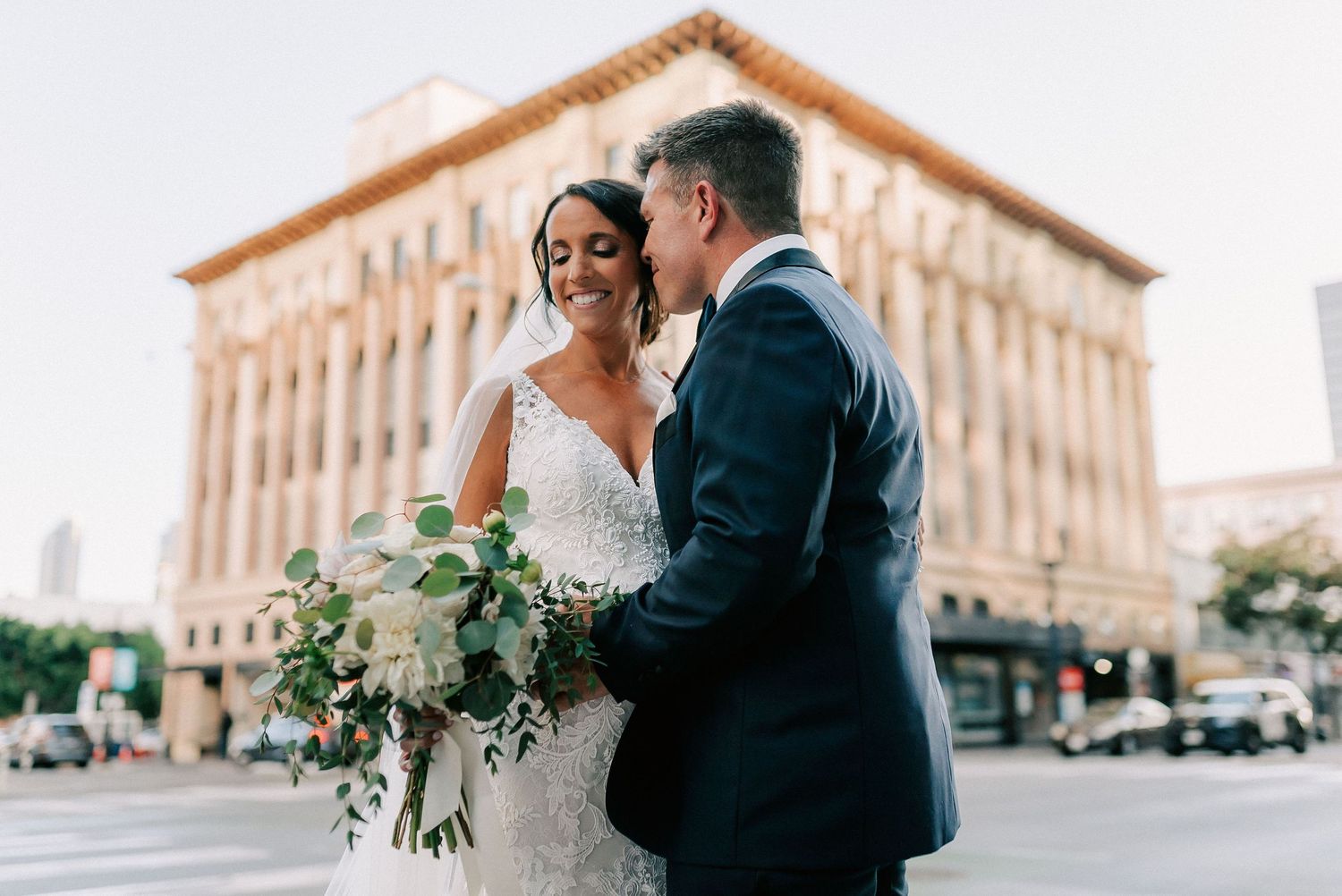 Bride and groom share a romantic moment outdoors, with bride holding white bouquet, in front of a classic building.