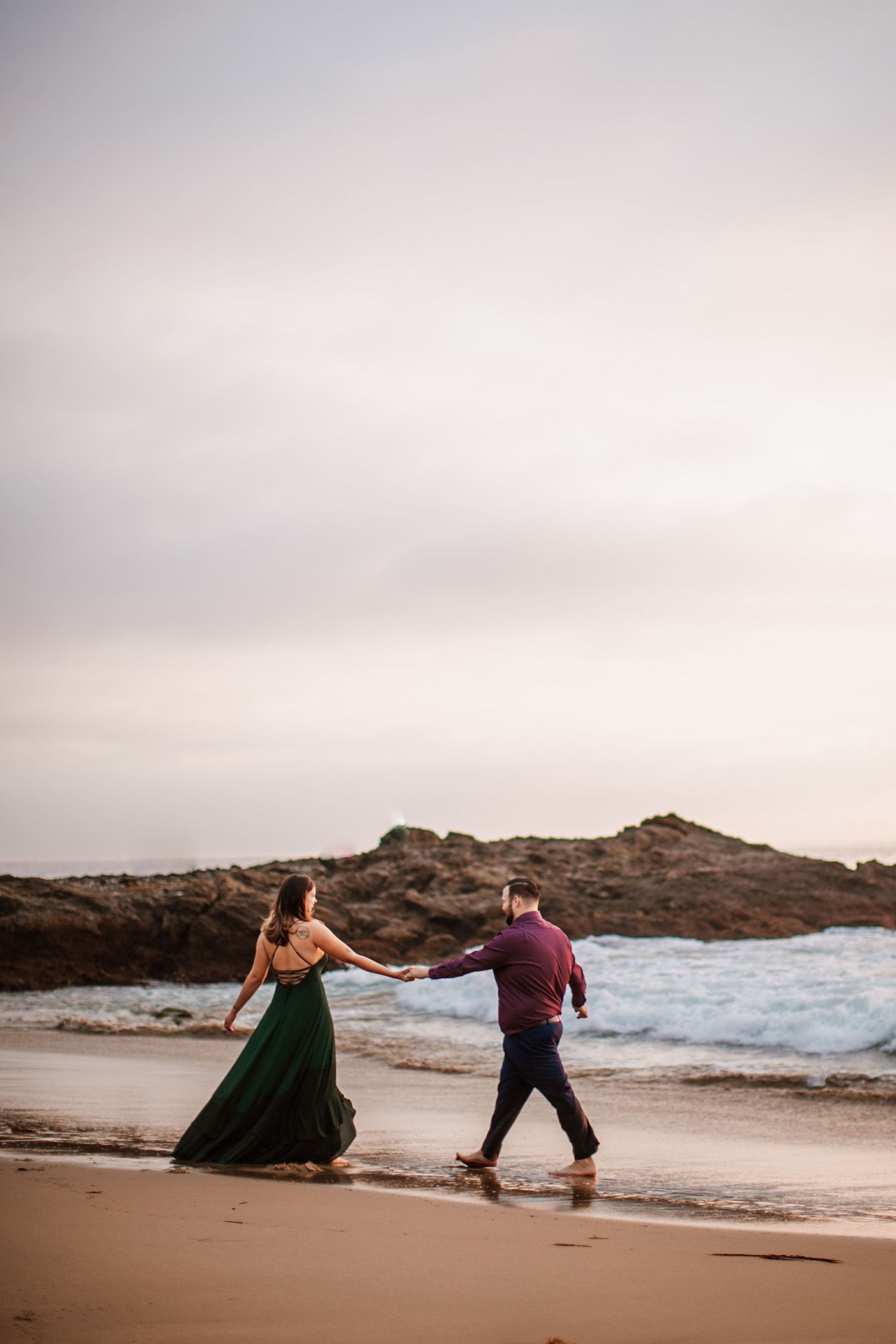 Couple holding hands walking on a sandy beach at sunset, woman in flowing green gown, man in burgundy shirt.