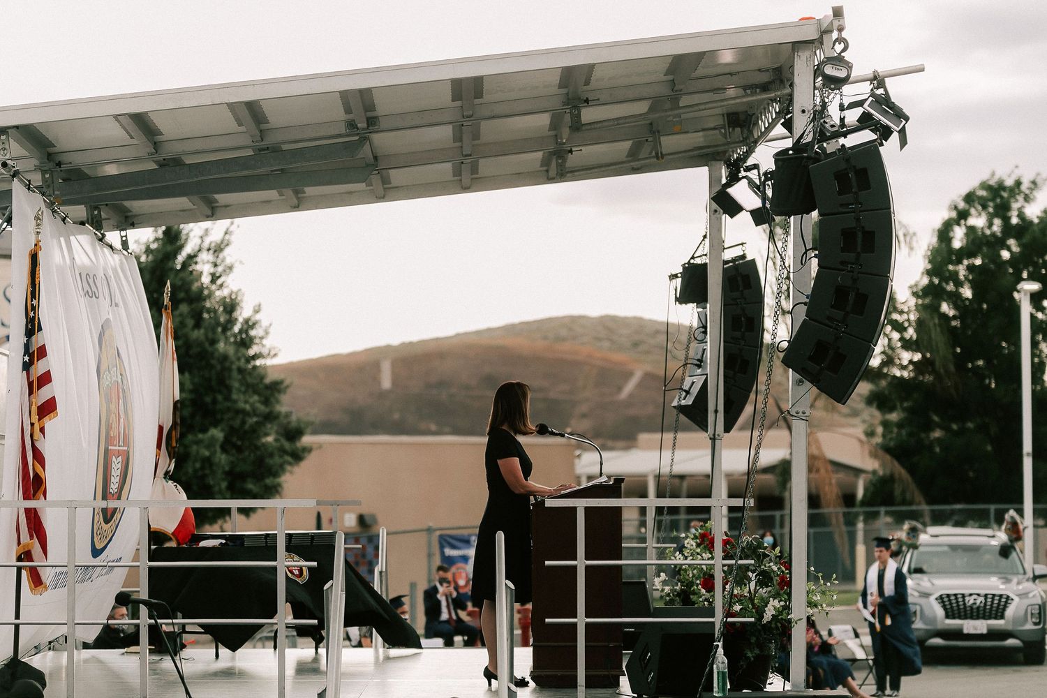 Woman speaking at an outdoor podium on a mobile stage with speakers, hills, and an American flag in the background.