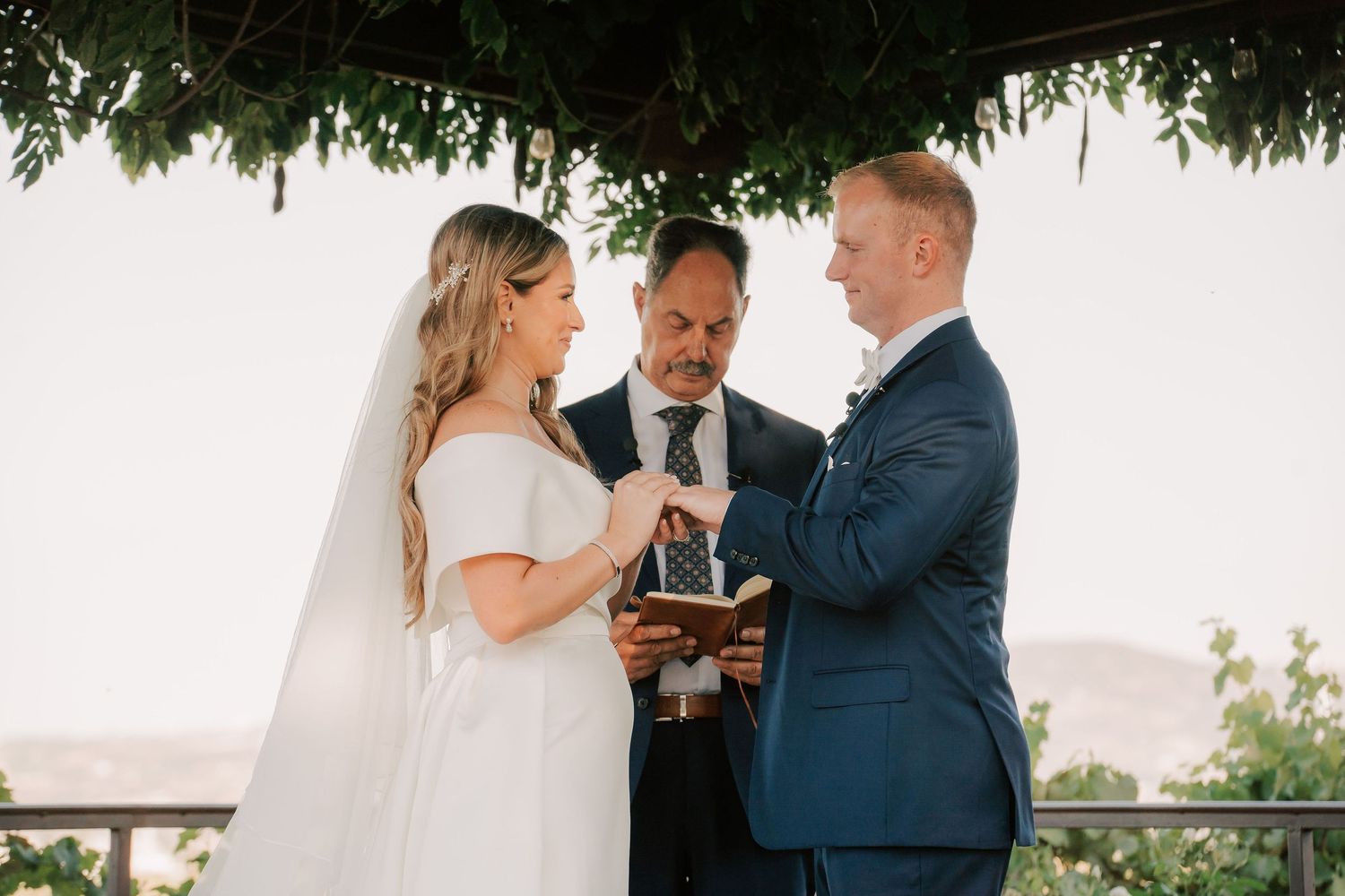 Bride and groom exchange vows under a floral arch with an officiant during an outdoor wedding ceremony.