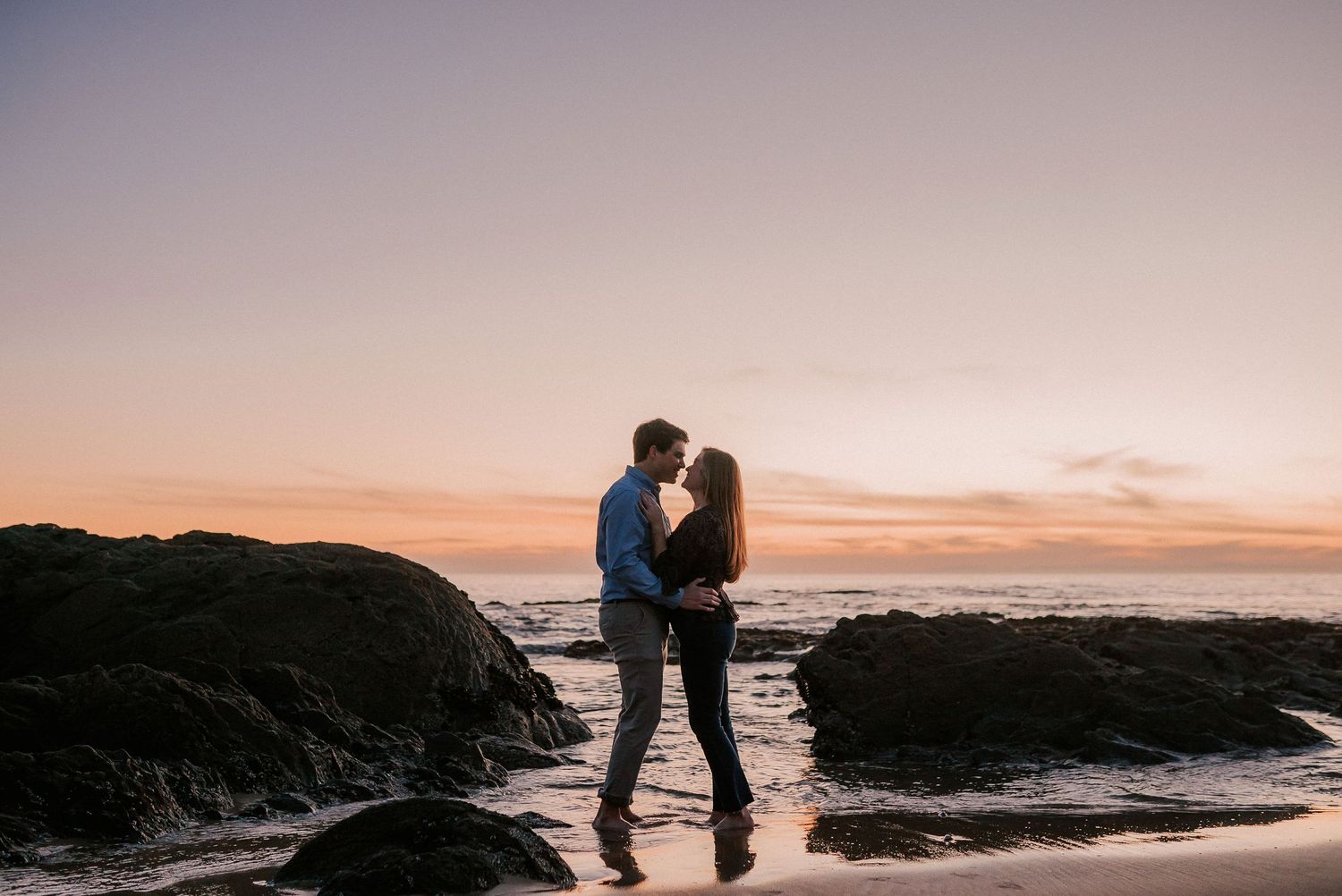 Couple sharing romantic moment near ocean rocks during stunning pink and orange sunset.