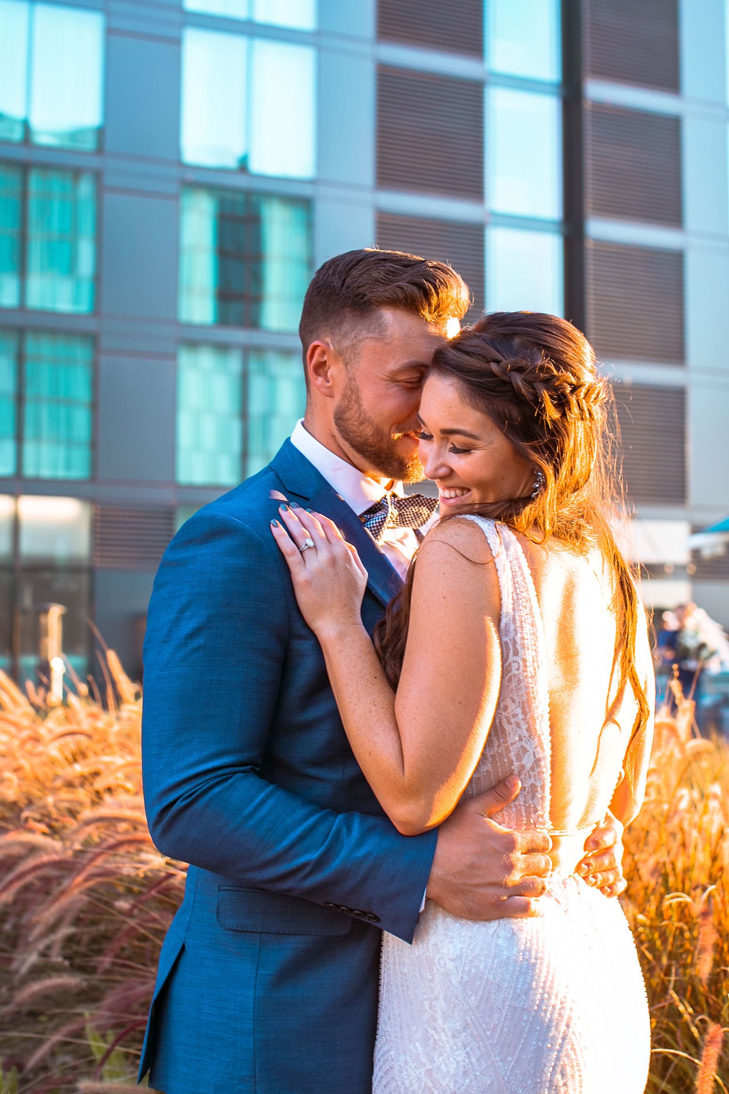 A couple in wedding attire embraces tenderly outdoors, golden sunlight glowing behind them near a modern building.