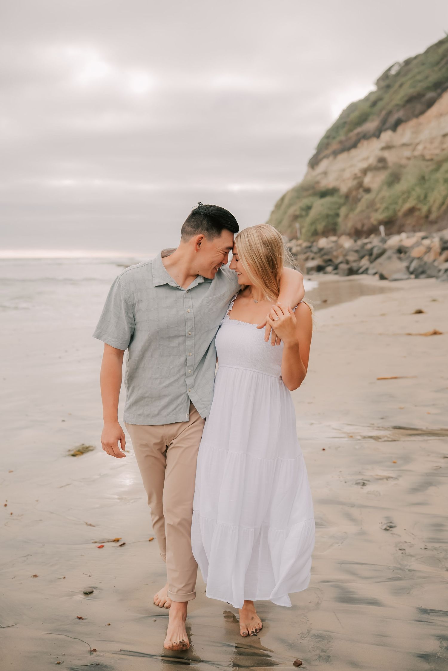 Couple walking barefoot on a sandy beach, woman in white dress and man in gray shirt, sharing a romantic moment near ocean cliffs.