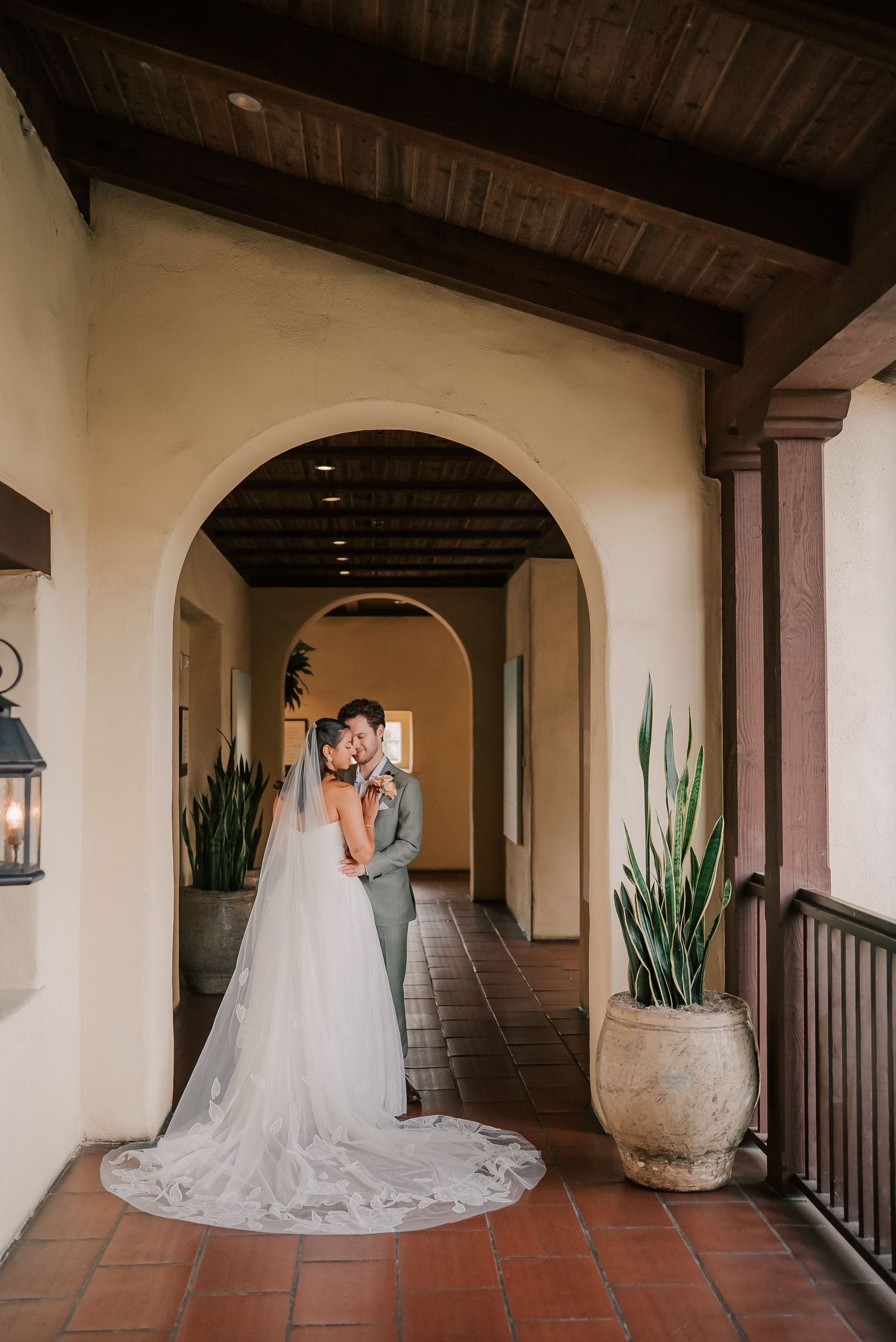 Bride and groom embrace under a Spanish-style archway, her flowing white gown trailing on terracotta tiles.