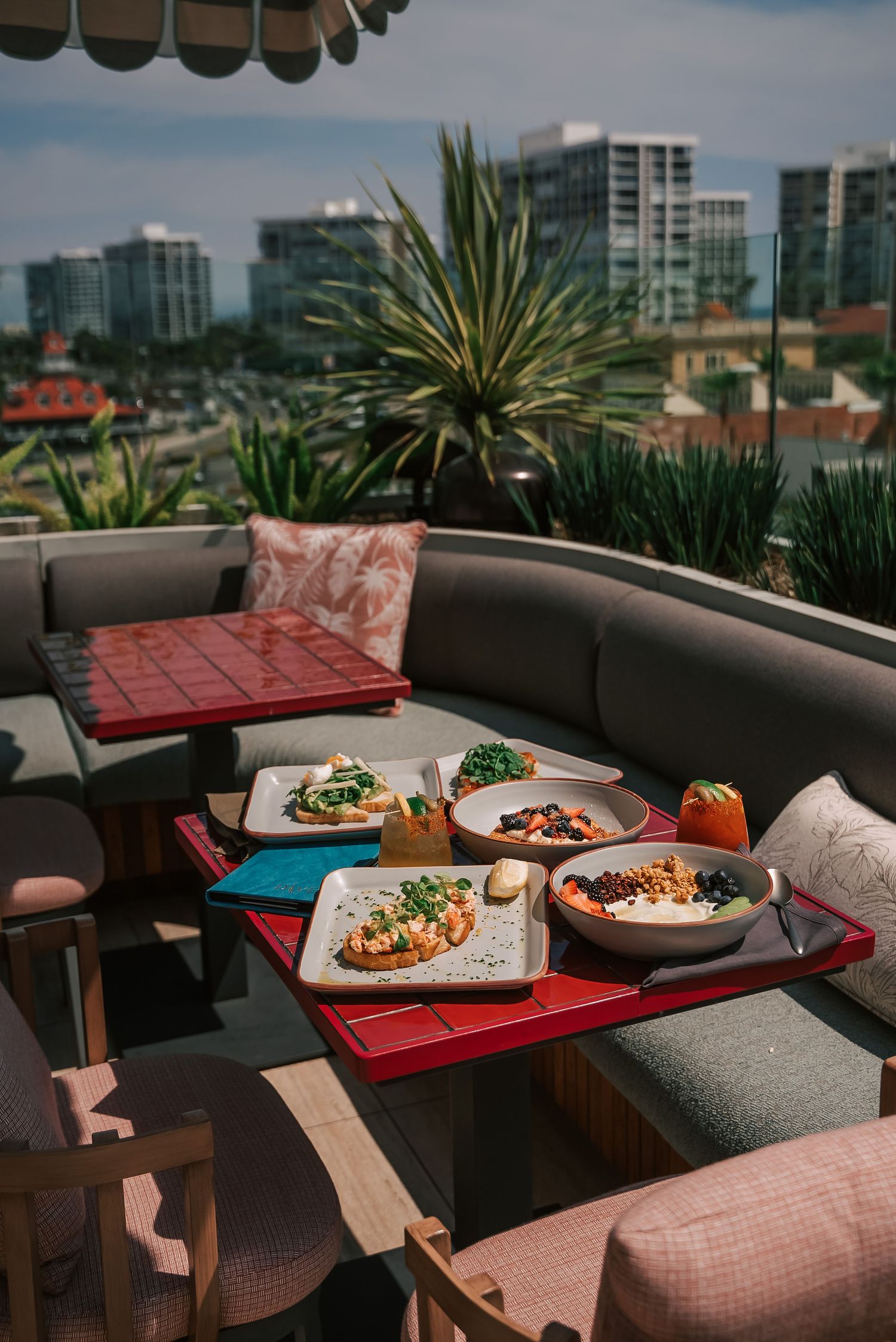 Rooftop dining spread with colorful dishes and drinks on a red tray, surrounded by lush plants and city skyline views.