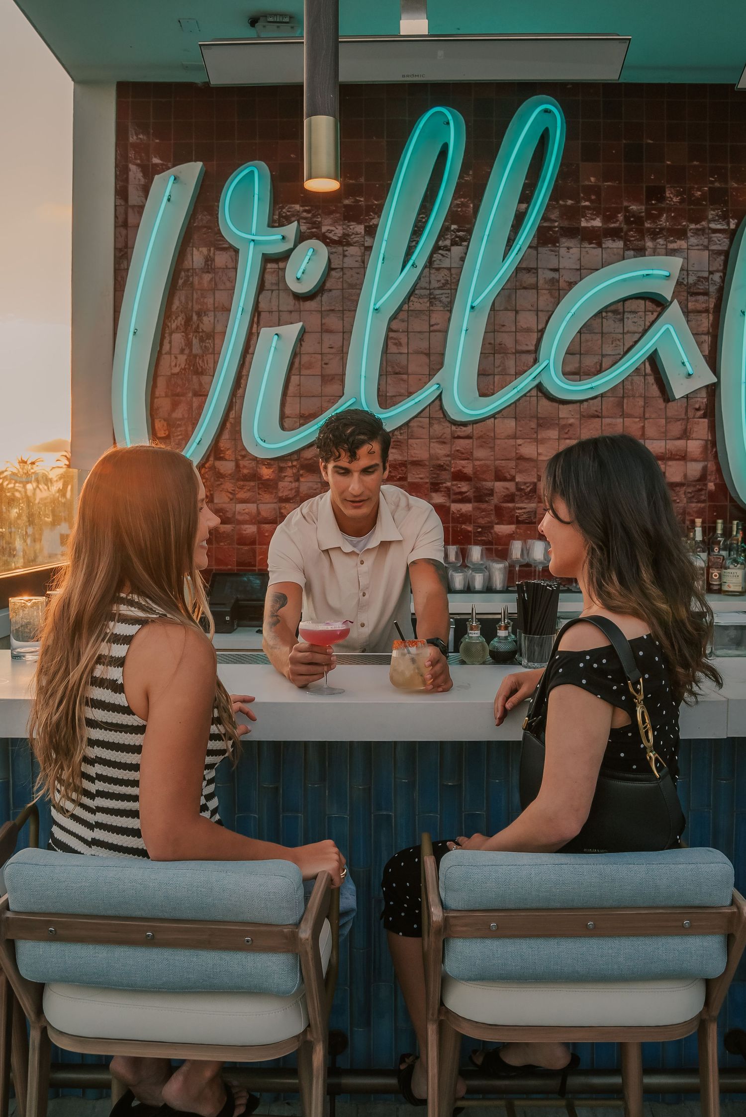 Two women sit at a rooftop bar called Villa, chatting with a male bartender holding a pink cocktail.
