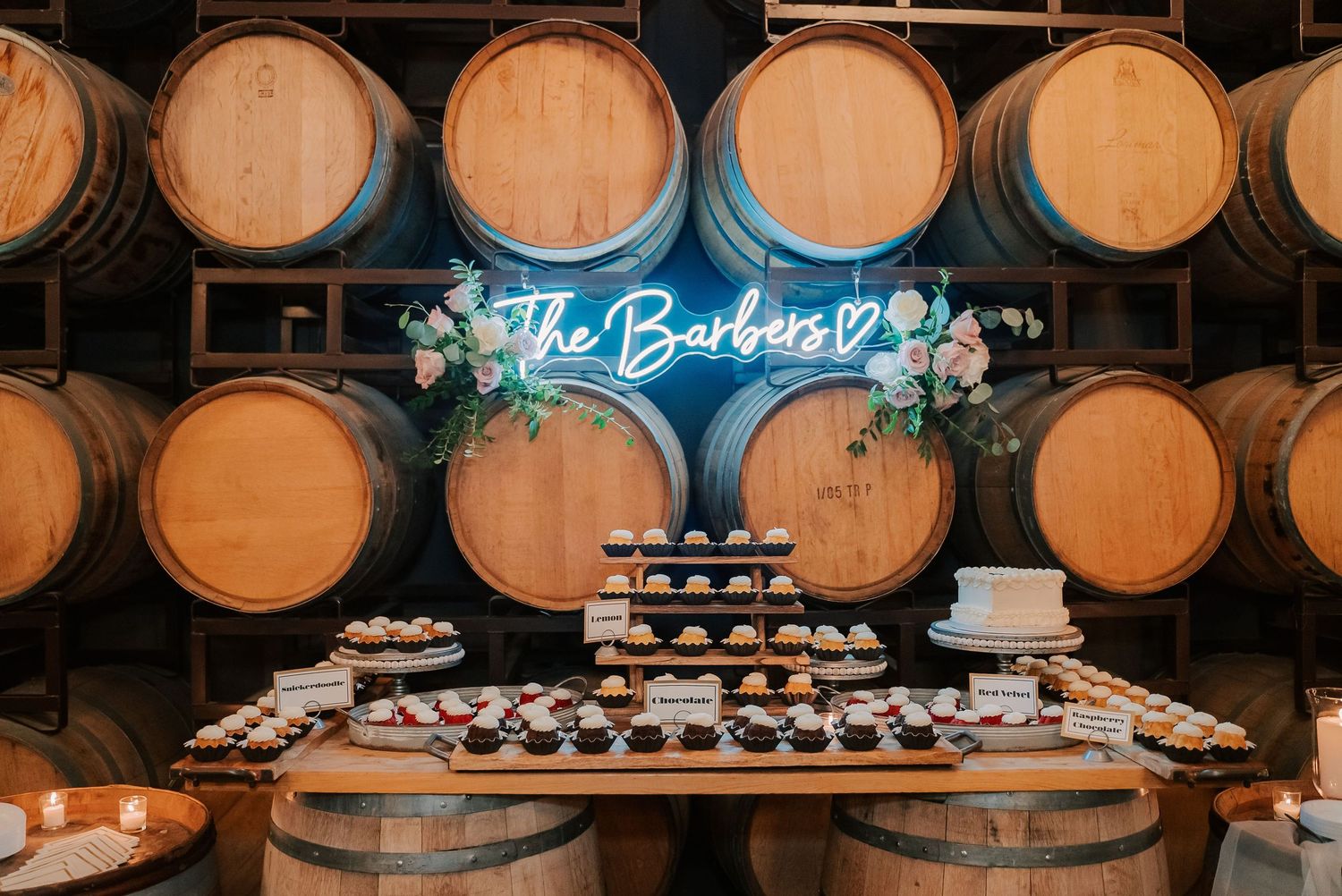 Wedding dessert table with cupcakes and cake displayed on barrels, neon sign reading The Barbers, floral decor.