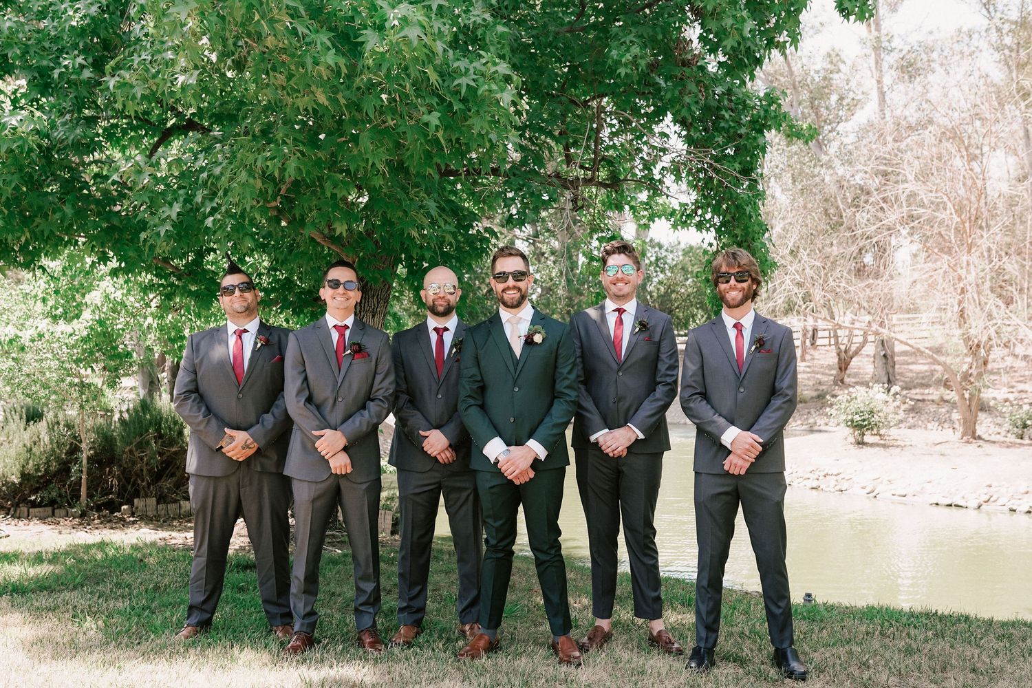 Groom in green suit stands with five groomsmen in gray suits and red ties outdoors near trees and a pond.