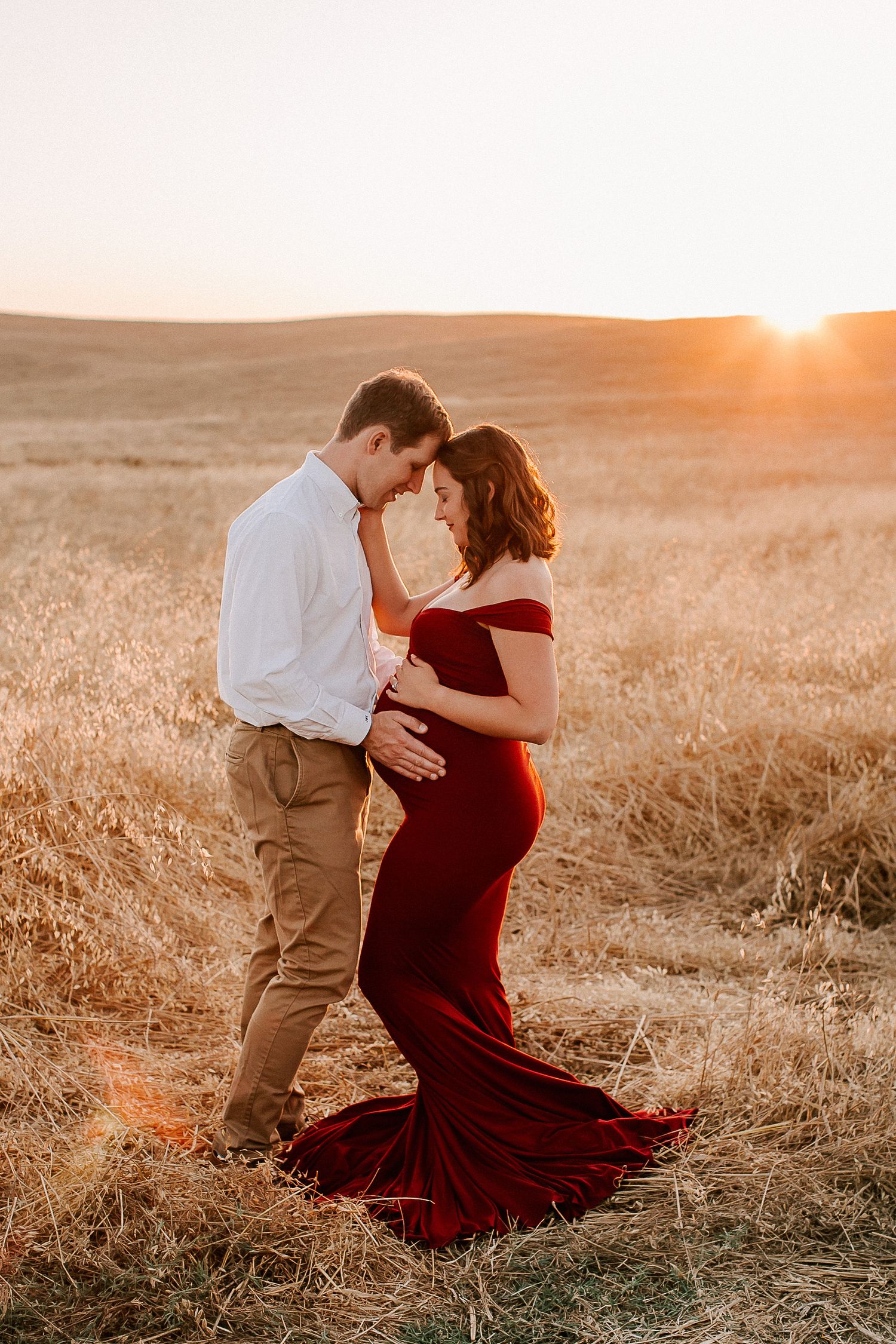 Couple sharing a tender moment in a golden field at sunset, woman in red maternity gown cradling her baby bump.