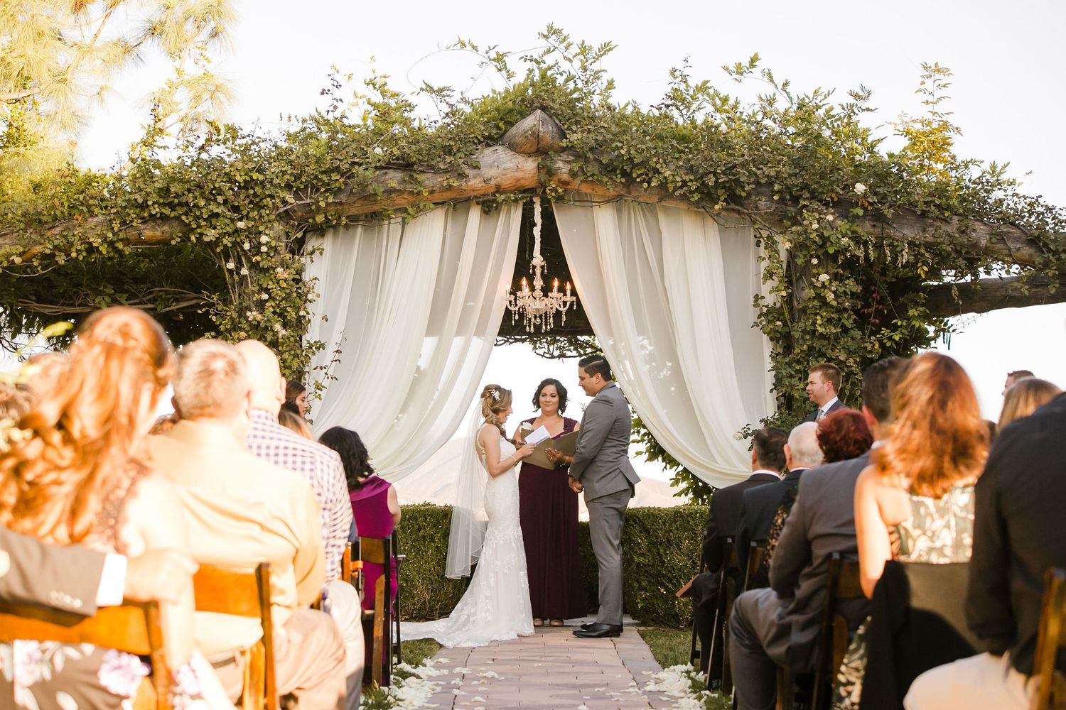 Outdoor wedding ceremony under a floral arch with white draping, guests seated on wooden chairs in a garden setting.
