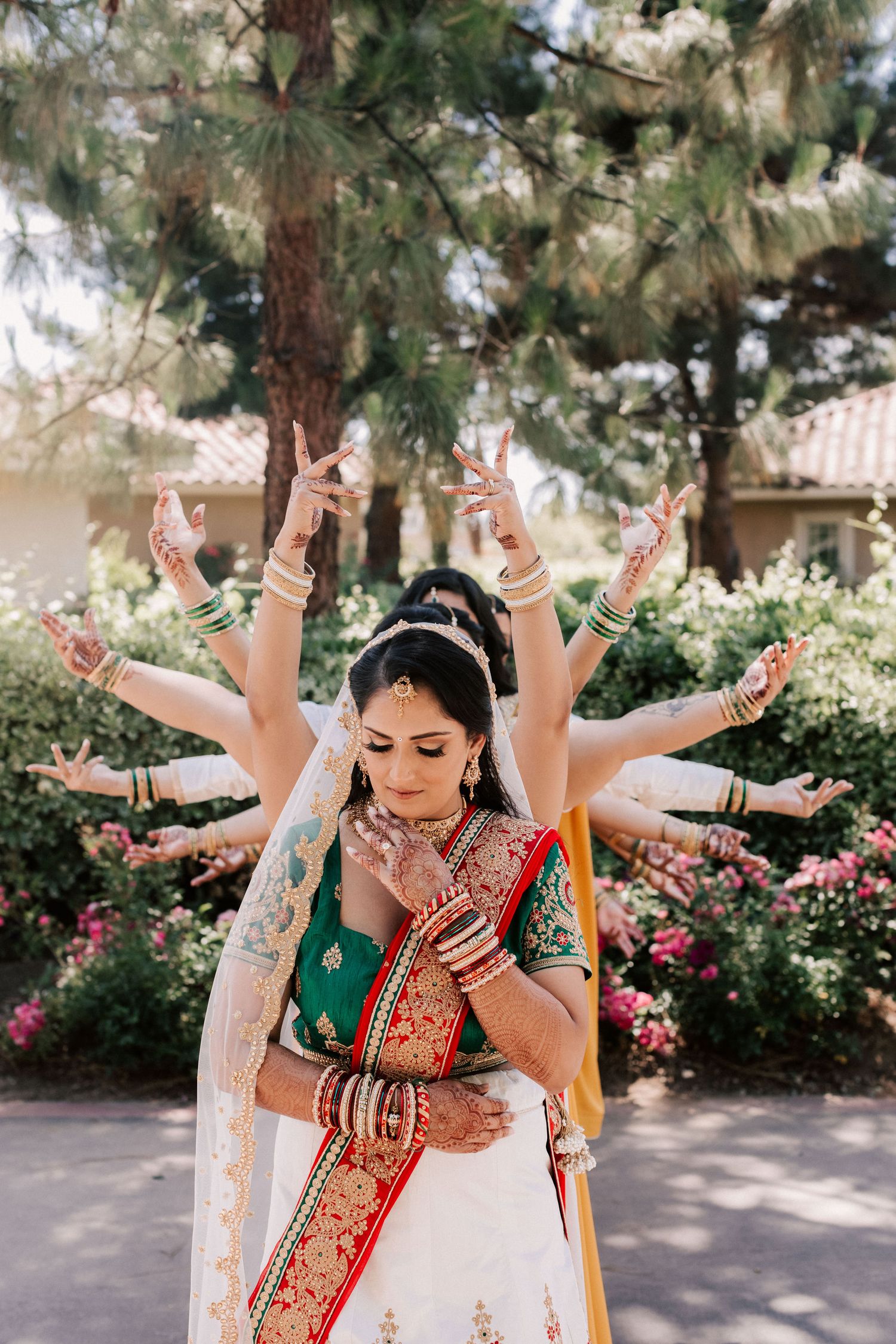 Indian bride in white and green lehenga poses with bridesmaids creating multi-arm goddess effect.