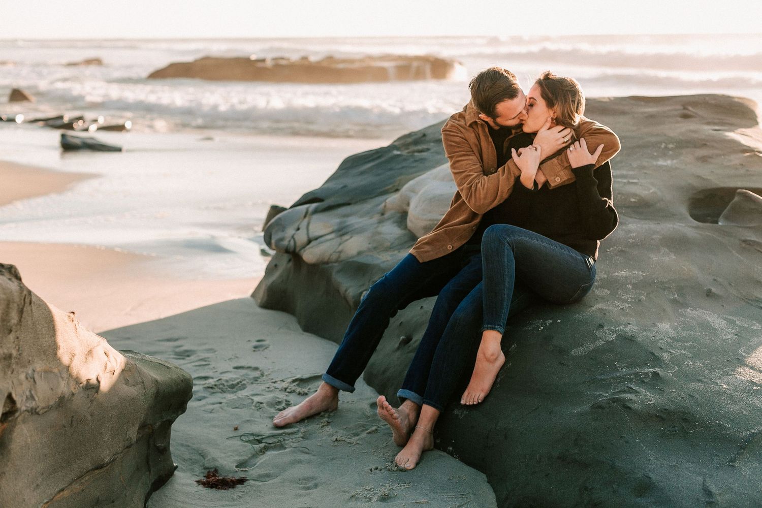 Couple sharing a romantic kiss while sitting together on large rocks at a scenic beach.