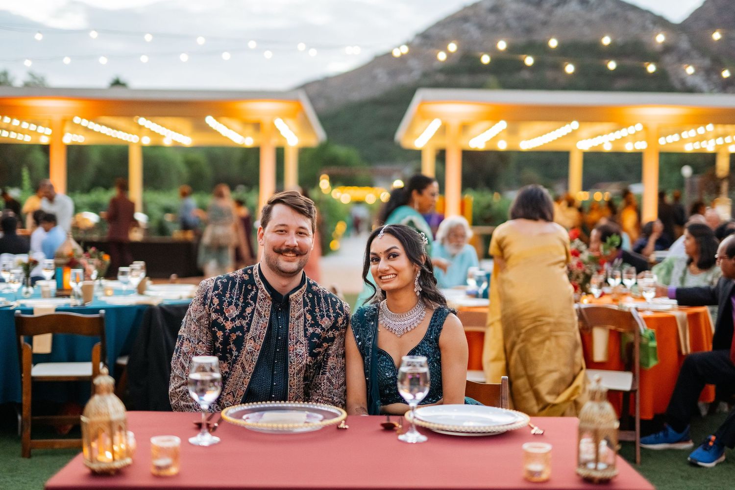 Couple smiling at outdoor wedding reception dinner table with string lights and mountain backdrop at dusk.