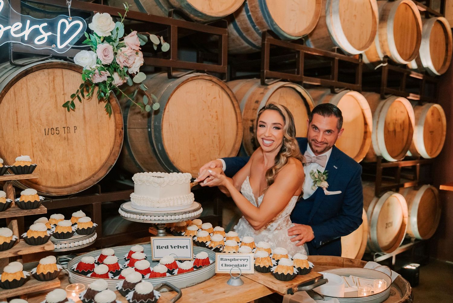 Bride and groom cutting wedding cake surrounded by cupcakes in a winery barrel room reception venue.