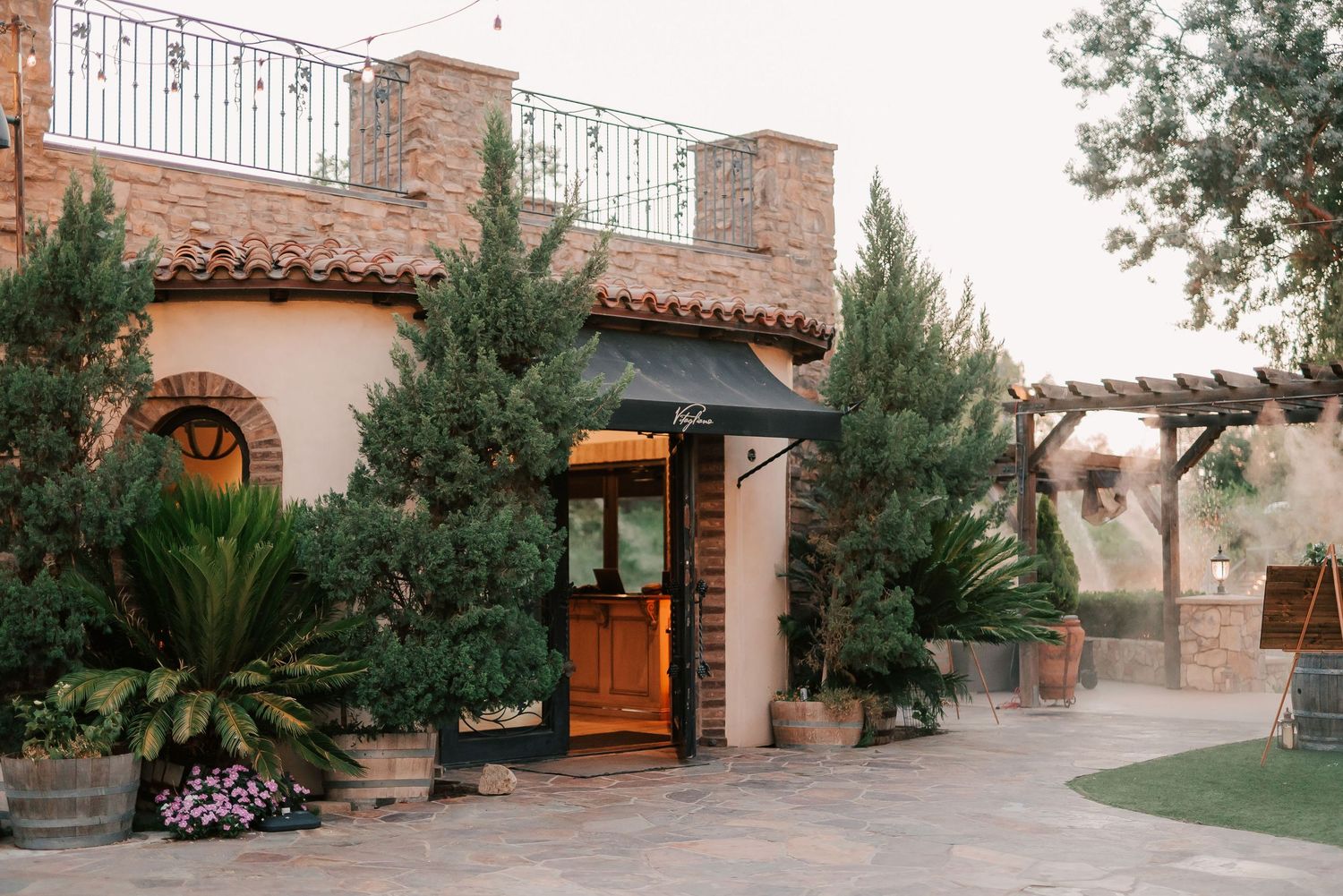Elegant Spanish-style venue entrance with wooden doors, lush greenery, and terracotta roof tiles at golden hour.