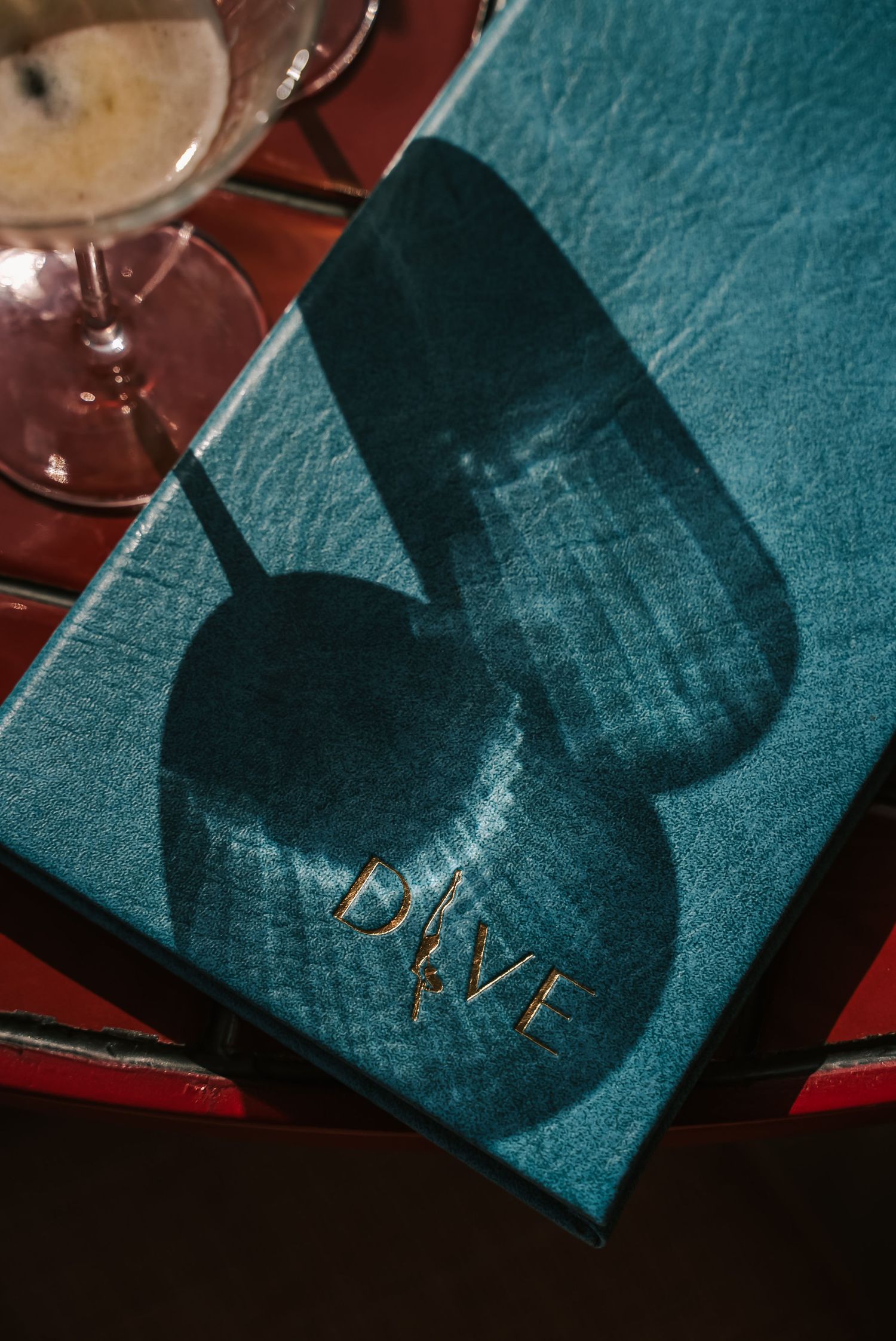 Teal leather menu with gold DIVE lettering on red table with wine glass shadow.