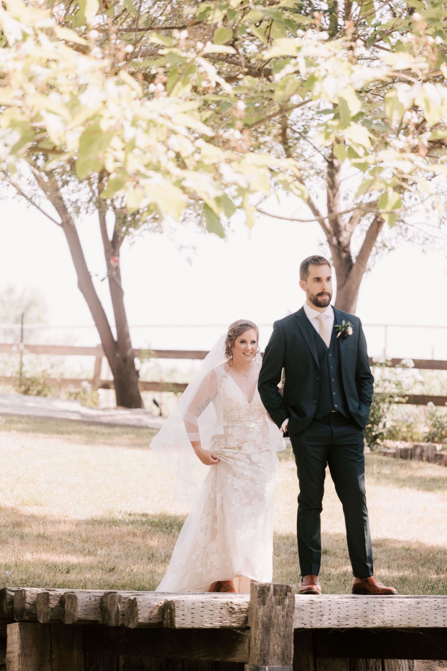 Bride in lace gown and groom in navy suit stand on wooden bridge surrounded by trees at outdoor wedding.