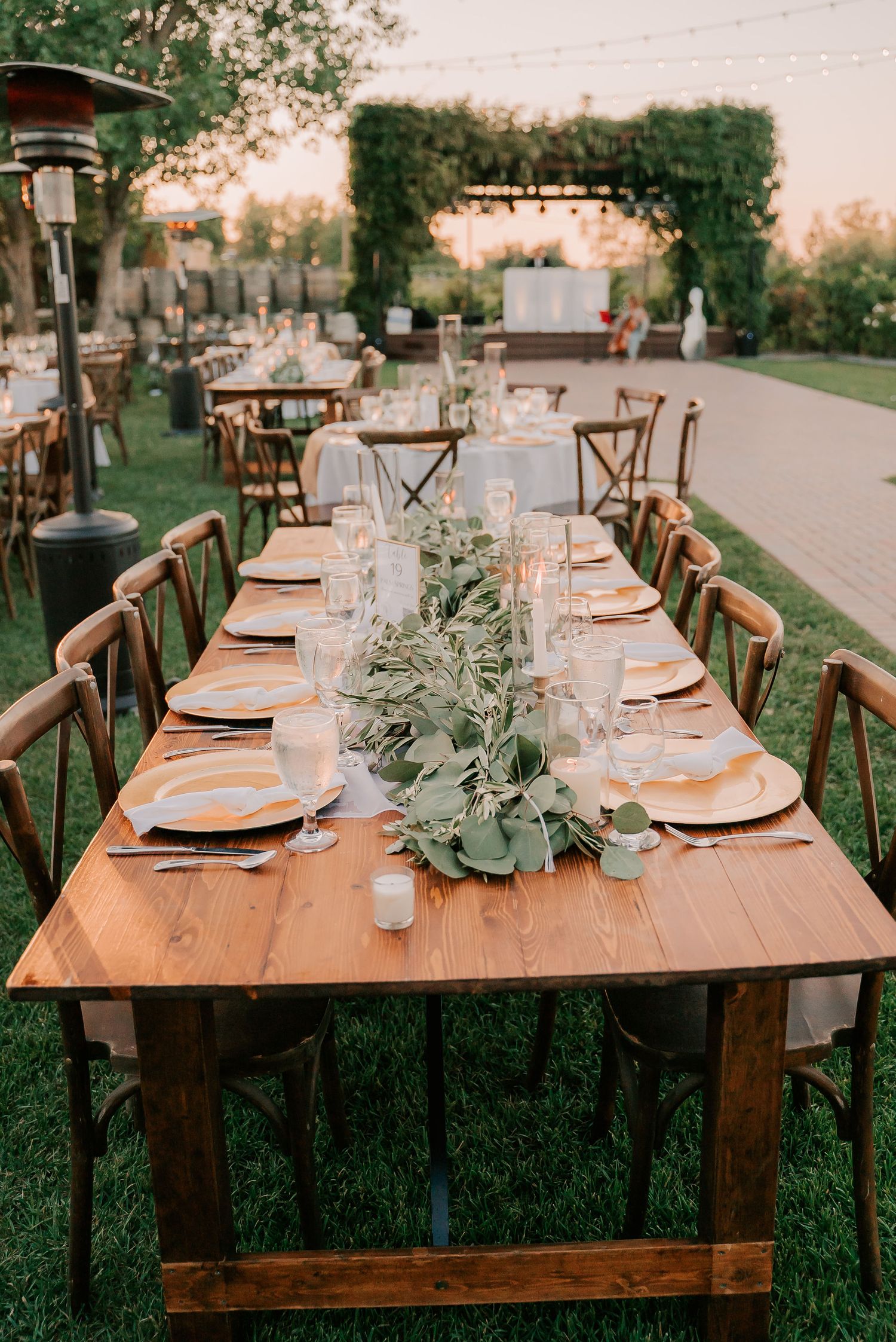 Rustic outdoor wedding reception with long wooden farm table, greenery runner, candles, and cross-back chairs at golden hour.