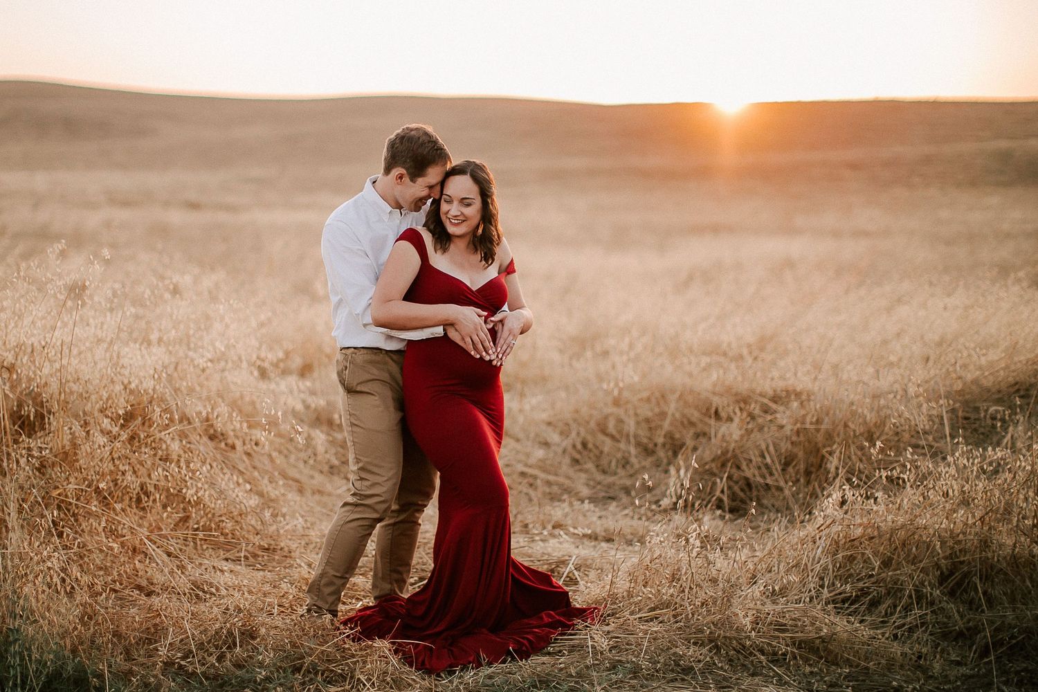 Couple embracing in golden field at sunset, woman in red maternity gown, romantic outdoor maternity photo session.