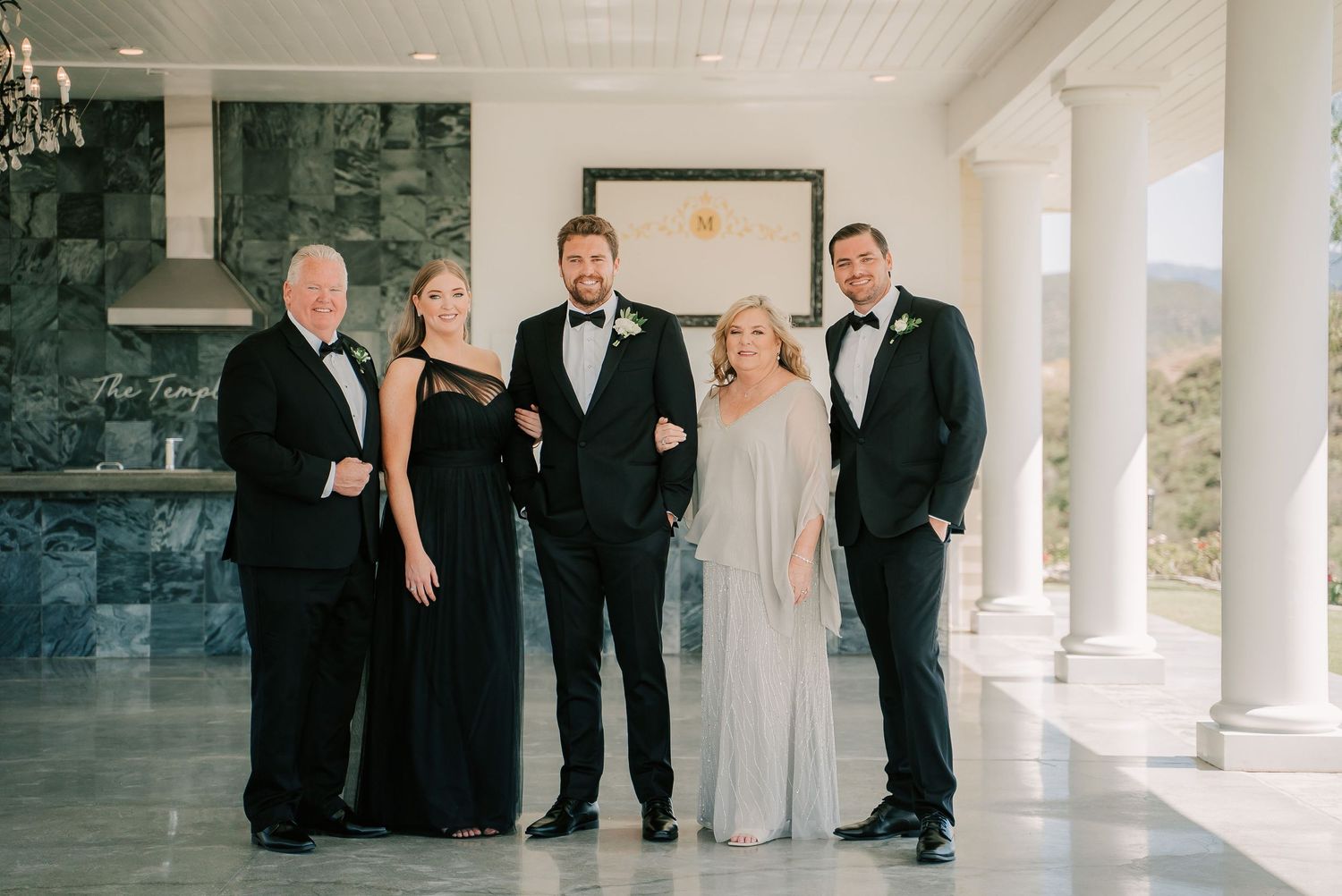 Elegant wedding family portrait with five guests in formal attire posing inside a venue with white columns.
