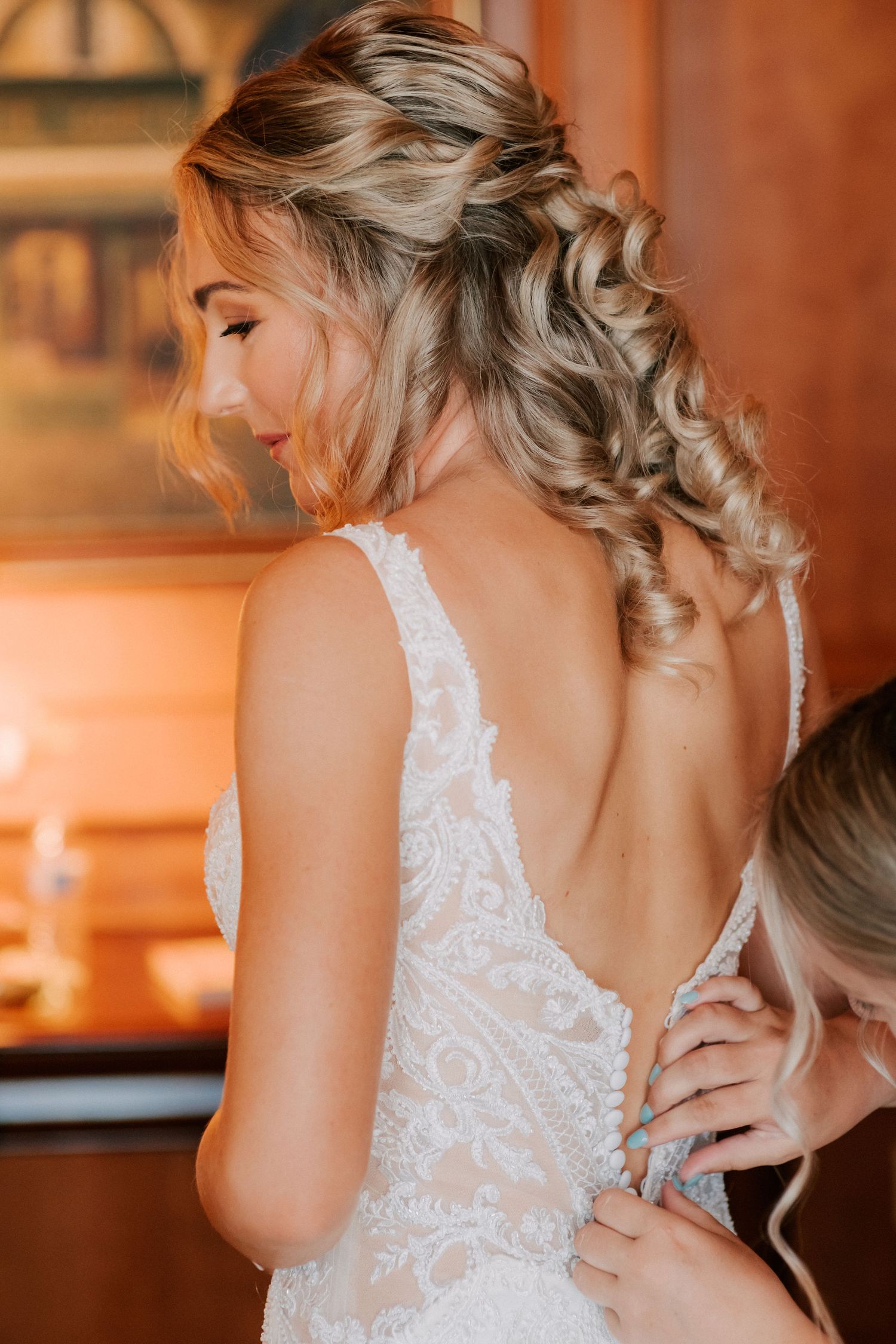 Bride in a backless lace wedding gown gets her dress fastened by a bridesmaid, showcasing an elegant updo hairstyle.