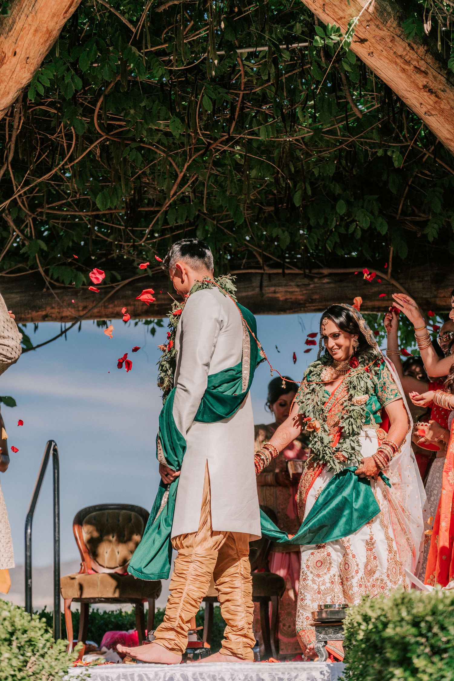 Bride and groom stand under floral arch as rose petals fall during Indian wedding ceremony.
