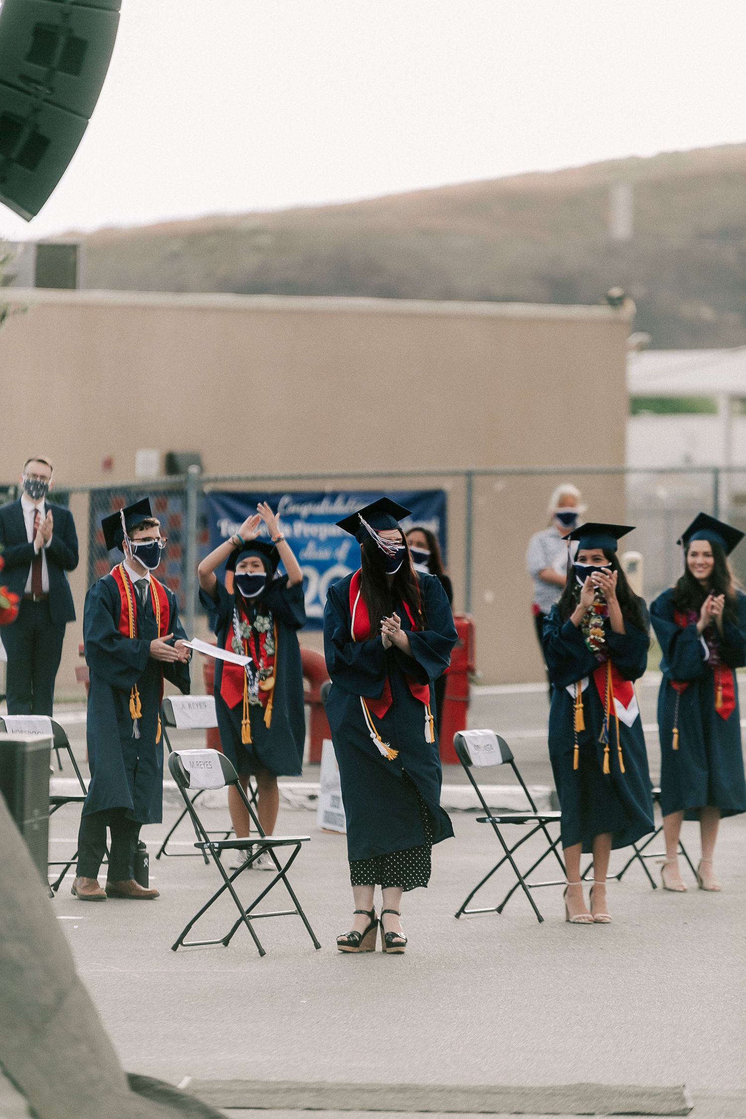 Graduates in black caps and gowns with red sashes celebrate outdoors at a socially distanced graduation ceremony.
