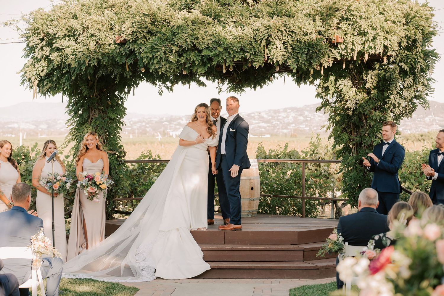 Bride and groom exchange vows under lush floral arch at outdoor wedding ceremony with scenic views.
