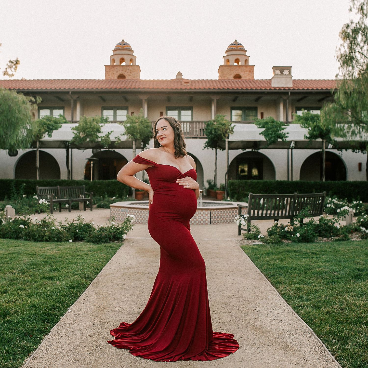 Pregnant woman in red off-shoulder mermaid gown poses in garden courtyard of elegant Spanish-style building.