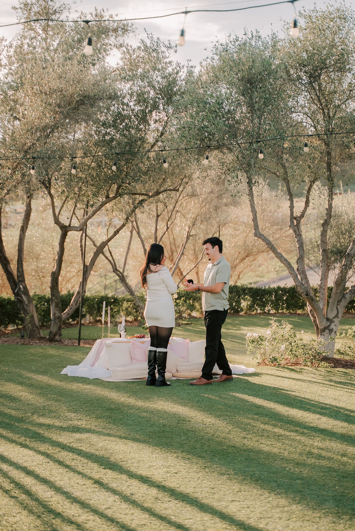 Couple sharing a romantic outdoor proposal moment beside a decorated table with candles and pink linens under olive trees.